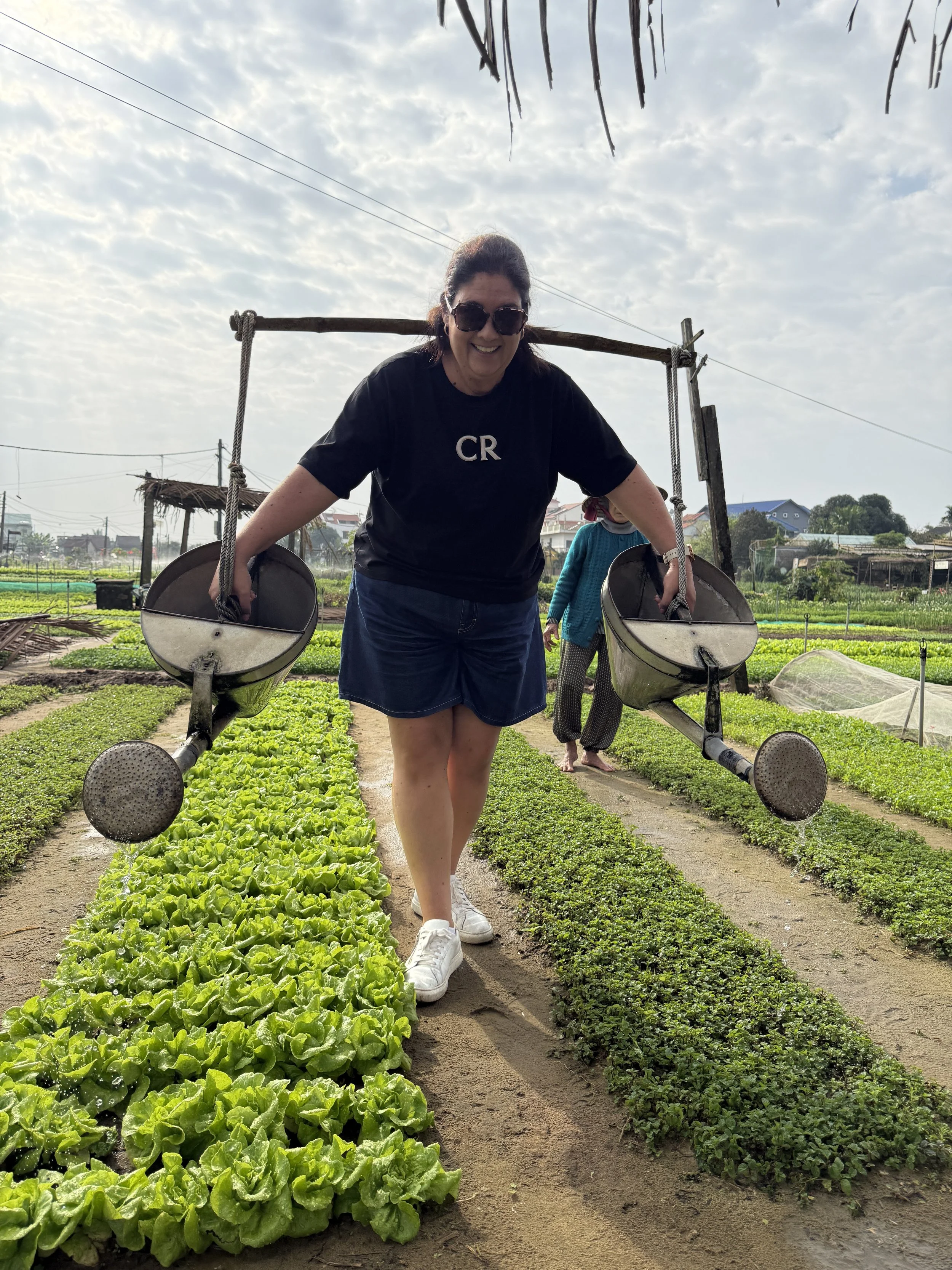 Woman smiling and carrying two watering cans in a farm field with rows of green lettuce, a person in the background, and sky with clouds overhead.