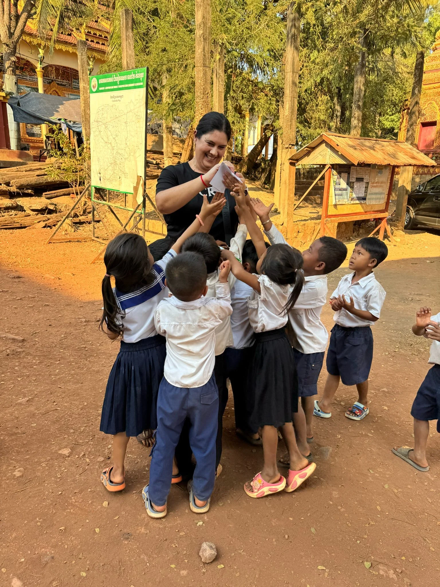 A group of young children are gathered around a woman outdoors, reaching out to touch or receive something from her. The children are dressed in uniforms and sandals, and the setting appears to be a rural or schoolyard area with trees, a signboard, a