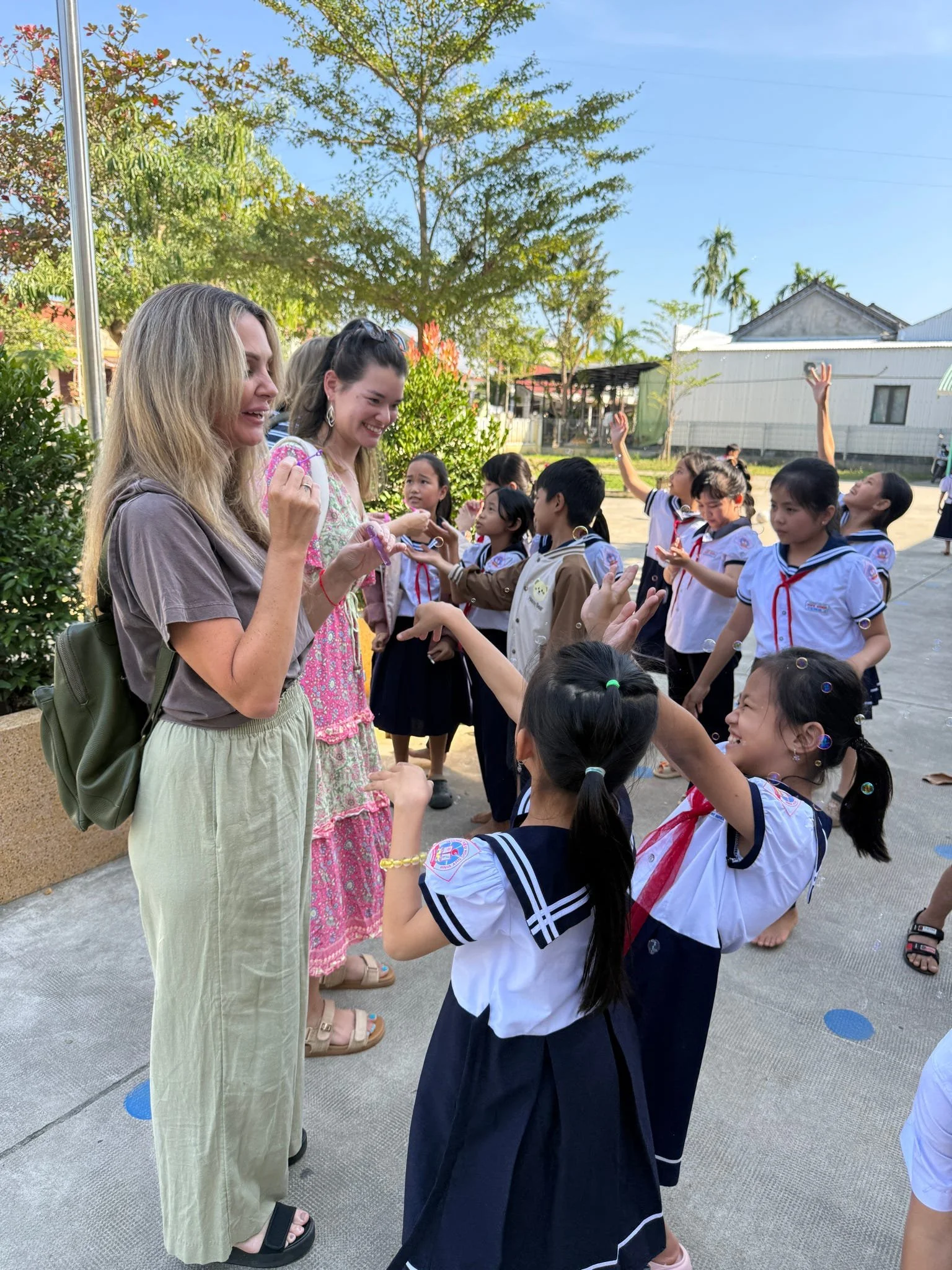 Two women are interacting with a group of children outdoors under a clear sky, with trees and buildings in the background. The children are wearing school uniforms and some are reaching out with their hands.