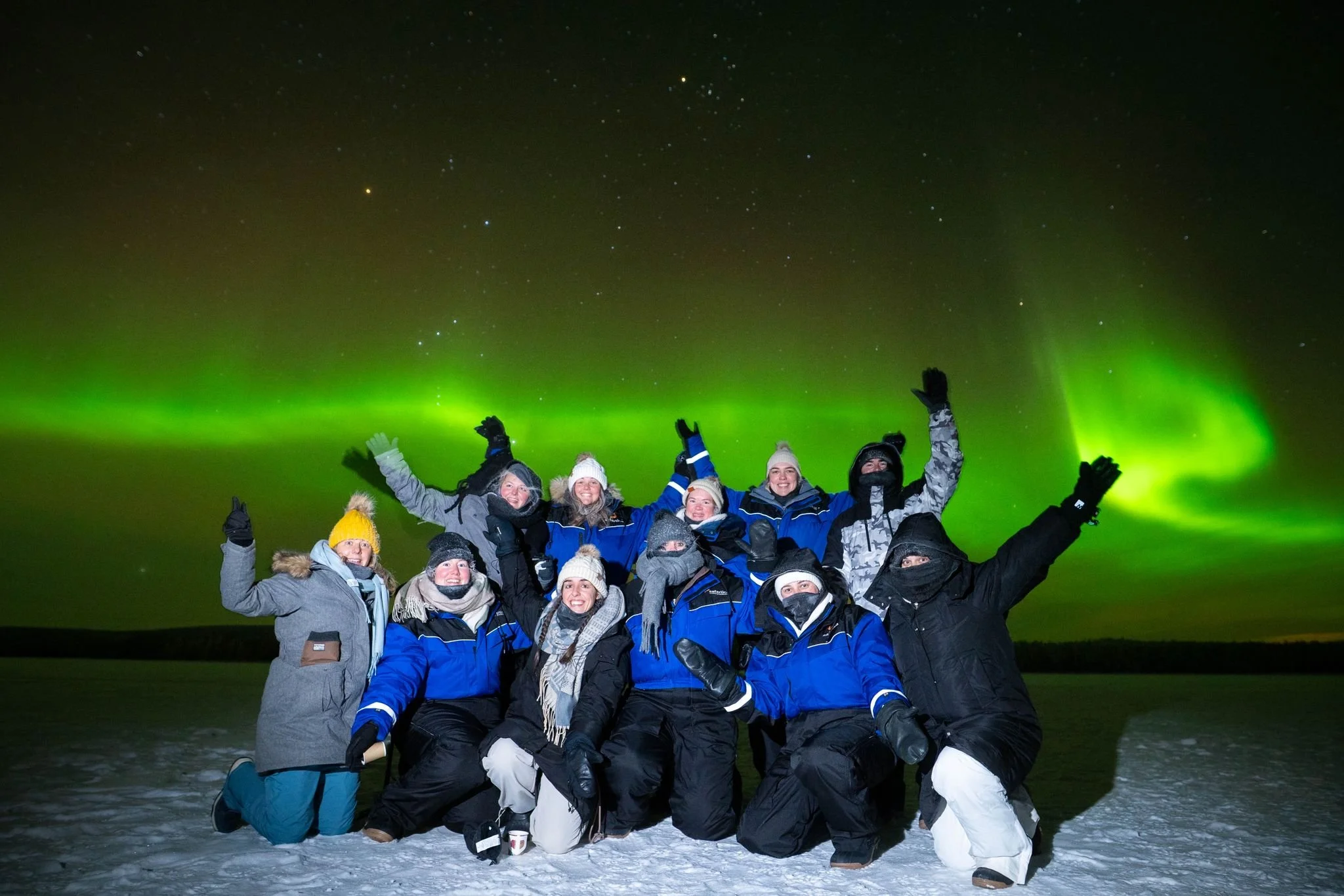 Group of people dressed in winter clothing posing outdoors at night under the northern lights.
