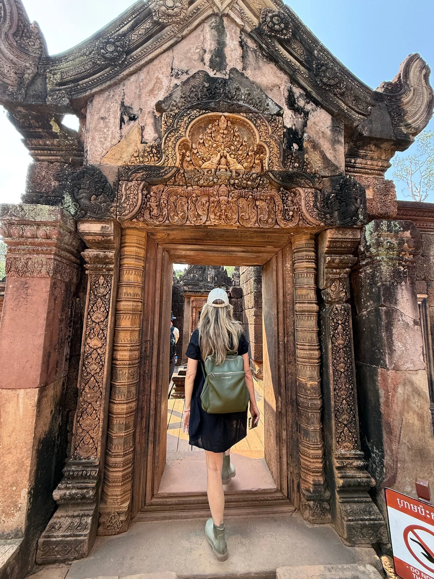 Woman with long hair, wearing a black dress, light green sneakers, and a gray backpack, walking through an ornate ancient stone doorway with intricate carvings, in a historical site, under a clear blue sky.