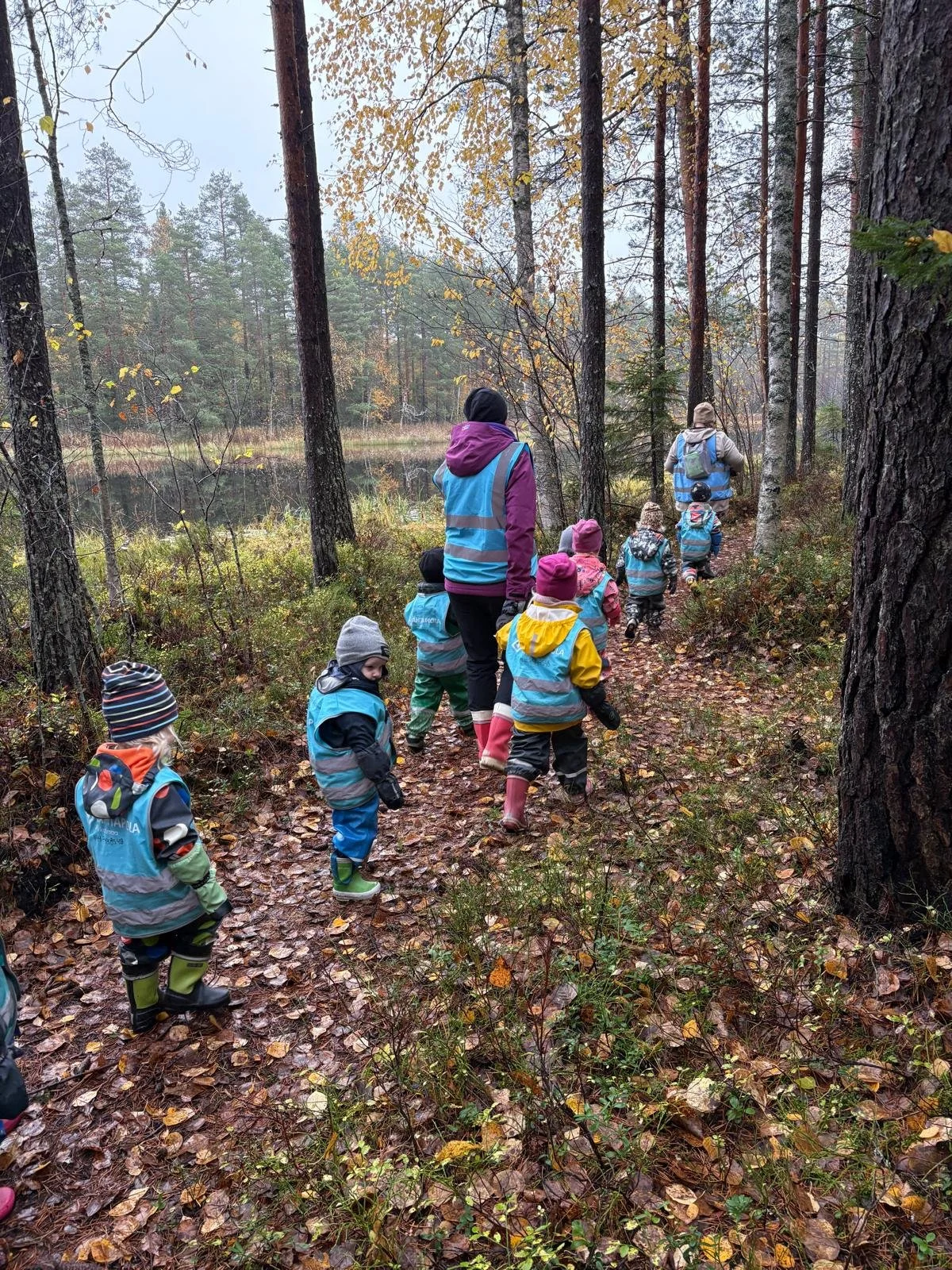 A group of children and two adults hiking through a forest trail during fall, with trees and a small lake in the background.