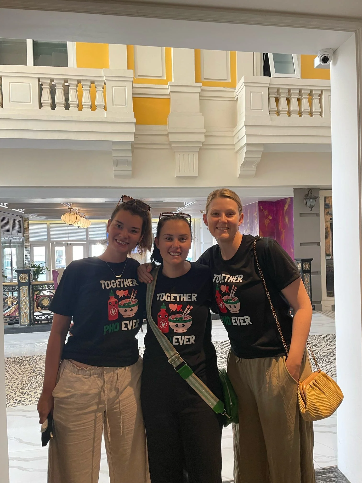 Three young women smiling and standing together inside a restaurant. They are wearing black T-shirts with colorful cartoon food items and the words 'TOGETHER PHO NEVER' printed on them. The restaurant has bright lighting, decorative walls, and a patt
