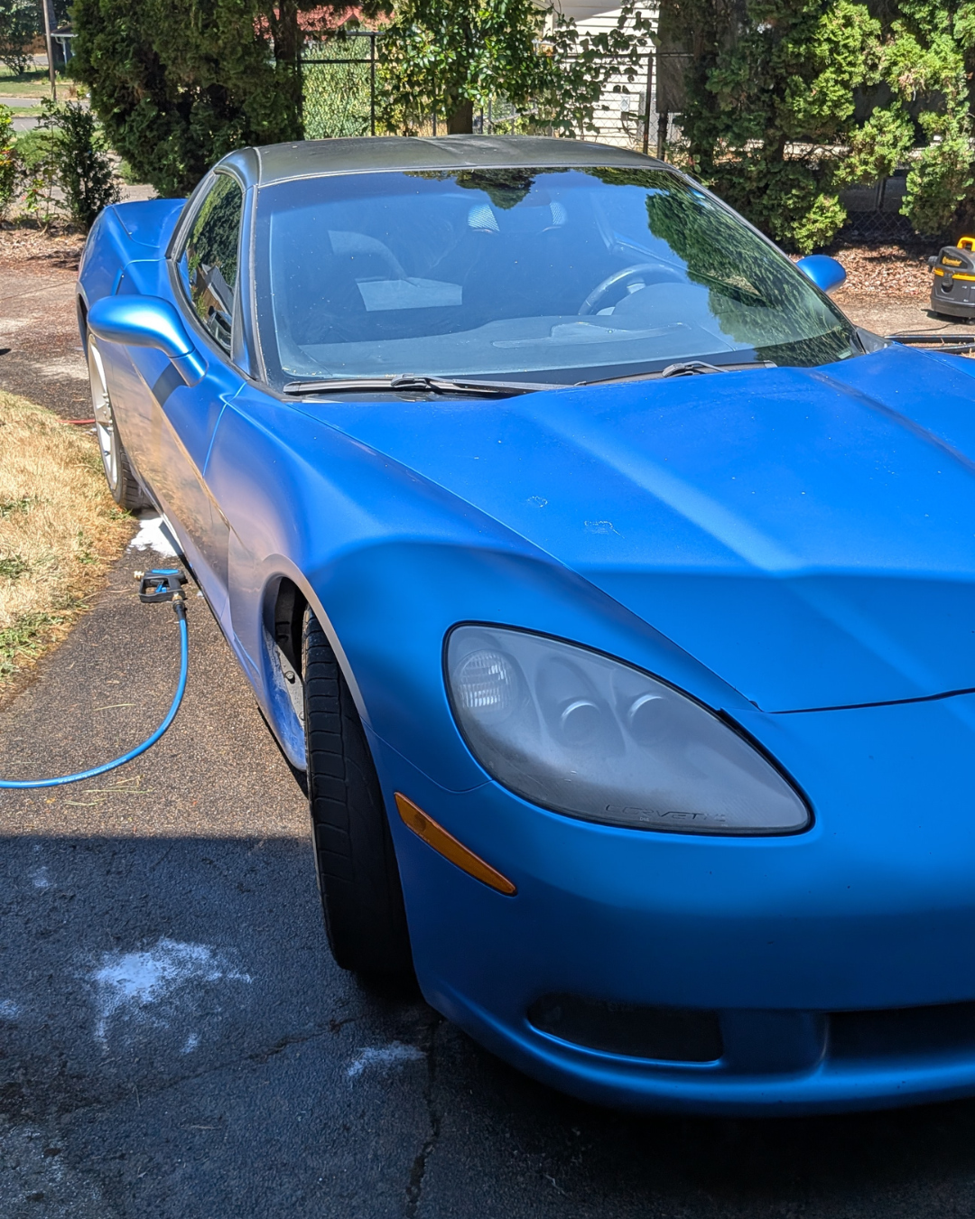 A blue Chevrolet Corvette sports car being washed outdoors on a driveway, connected to a hose for rinsing.