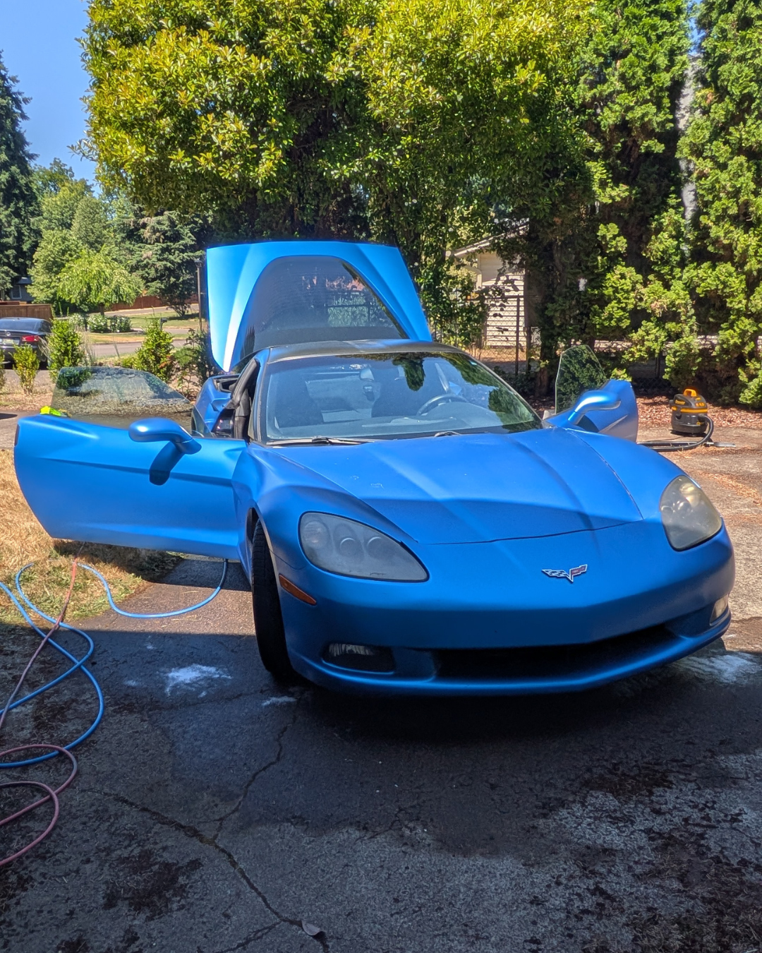 A blue Chevrolet Corvette with its doors open and hood raised, parked on a driveway with trees and a house in the background, during a sunny day.