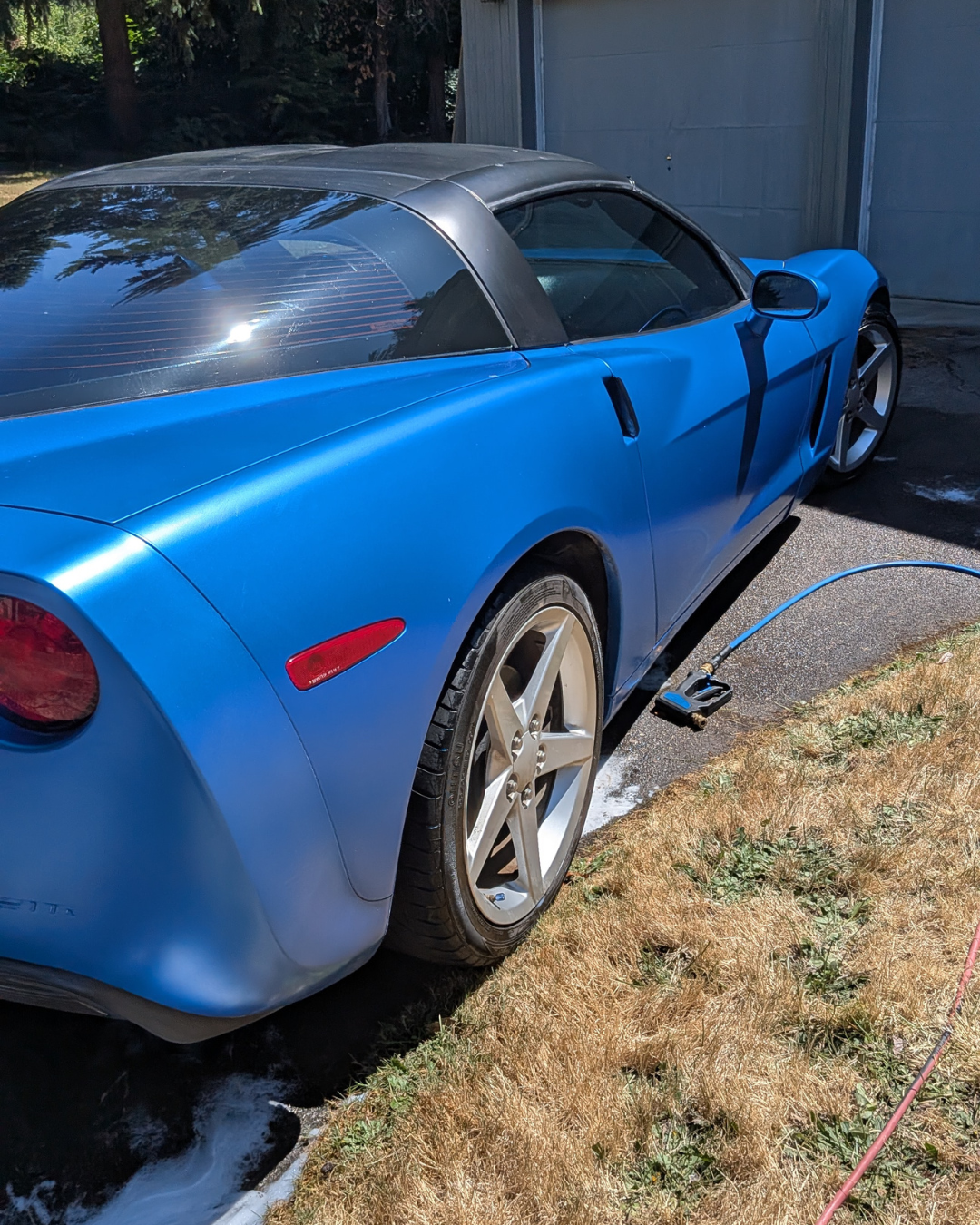 Blue Chevrolet Corvette sports car being washed outside with a hose on a concrete driveway.