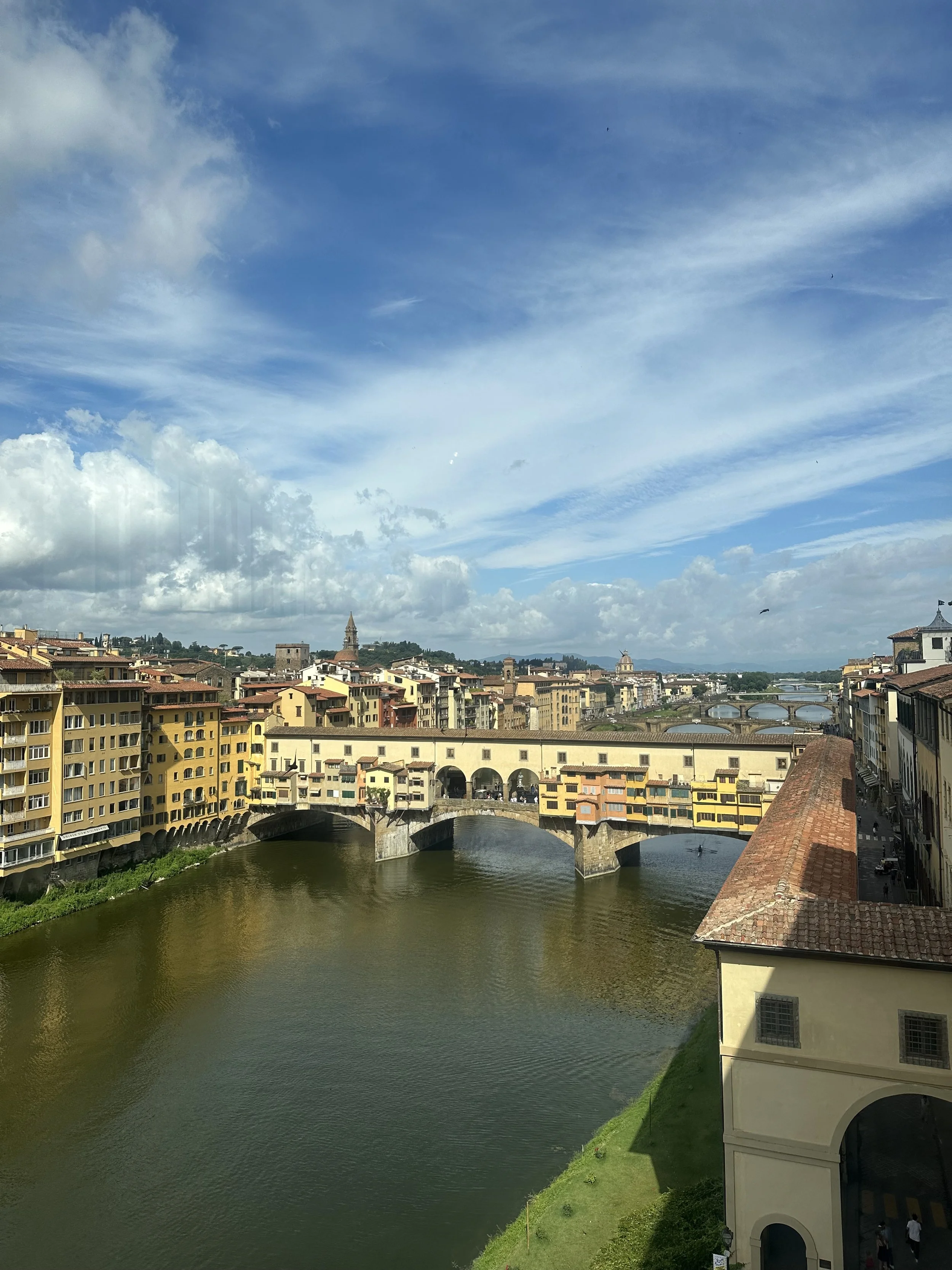 View of a historic Italian city with colorful buildings along a river, a stone bridge, and a cloudy blue sky.