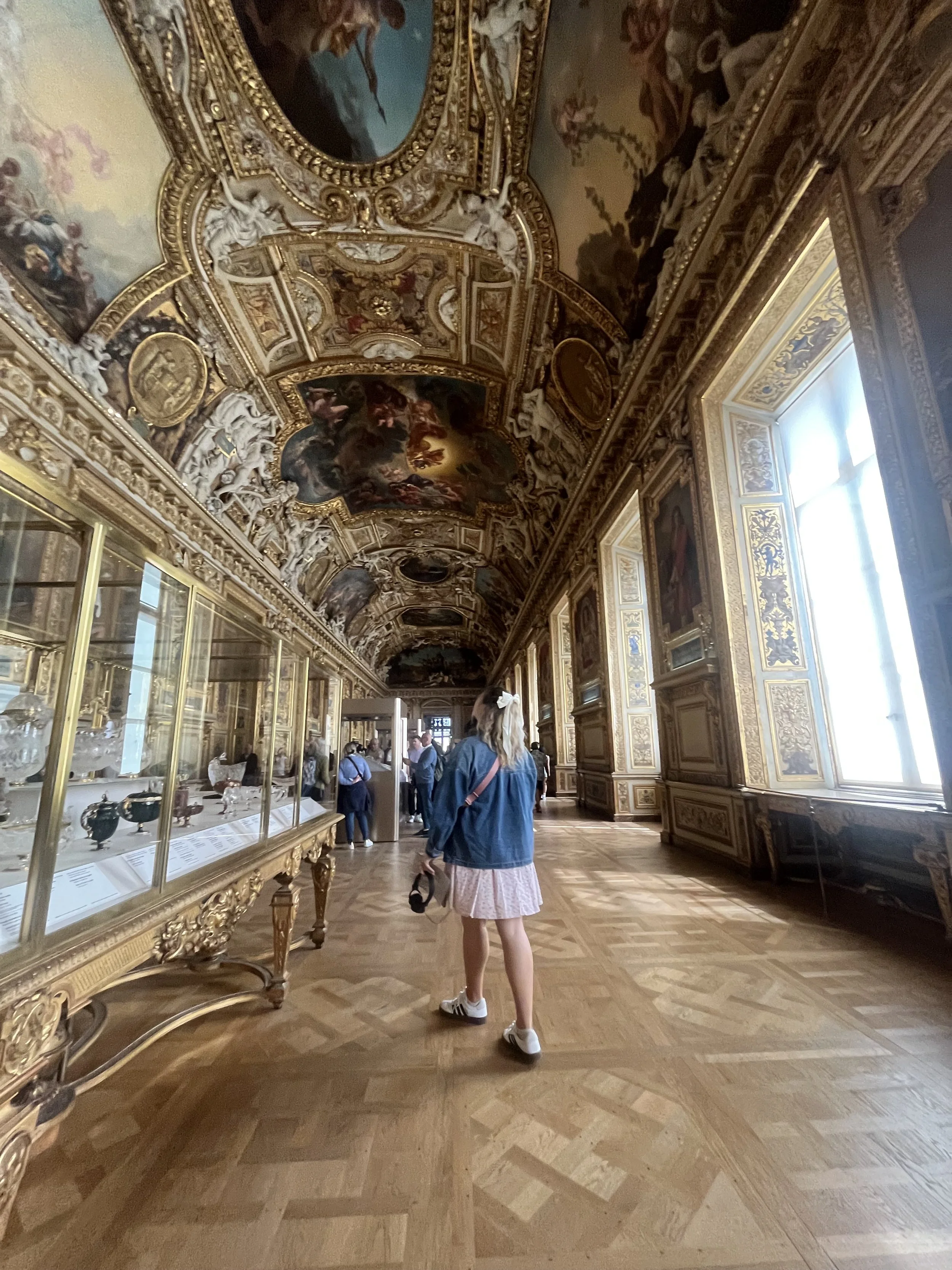 People inside an ornate room with gold-trimmed ceilings and walls, viewing artwork and artifacts, with a woman in the foreground wearing a light pink skirt, a denim jacket, and sneakers.