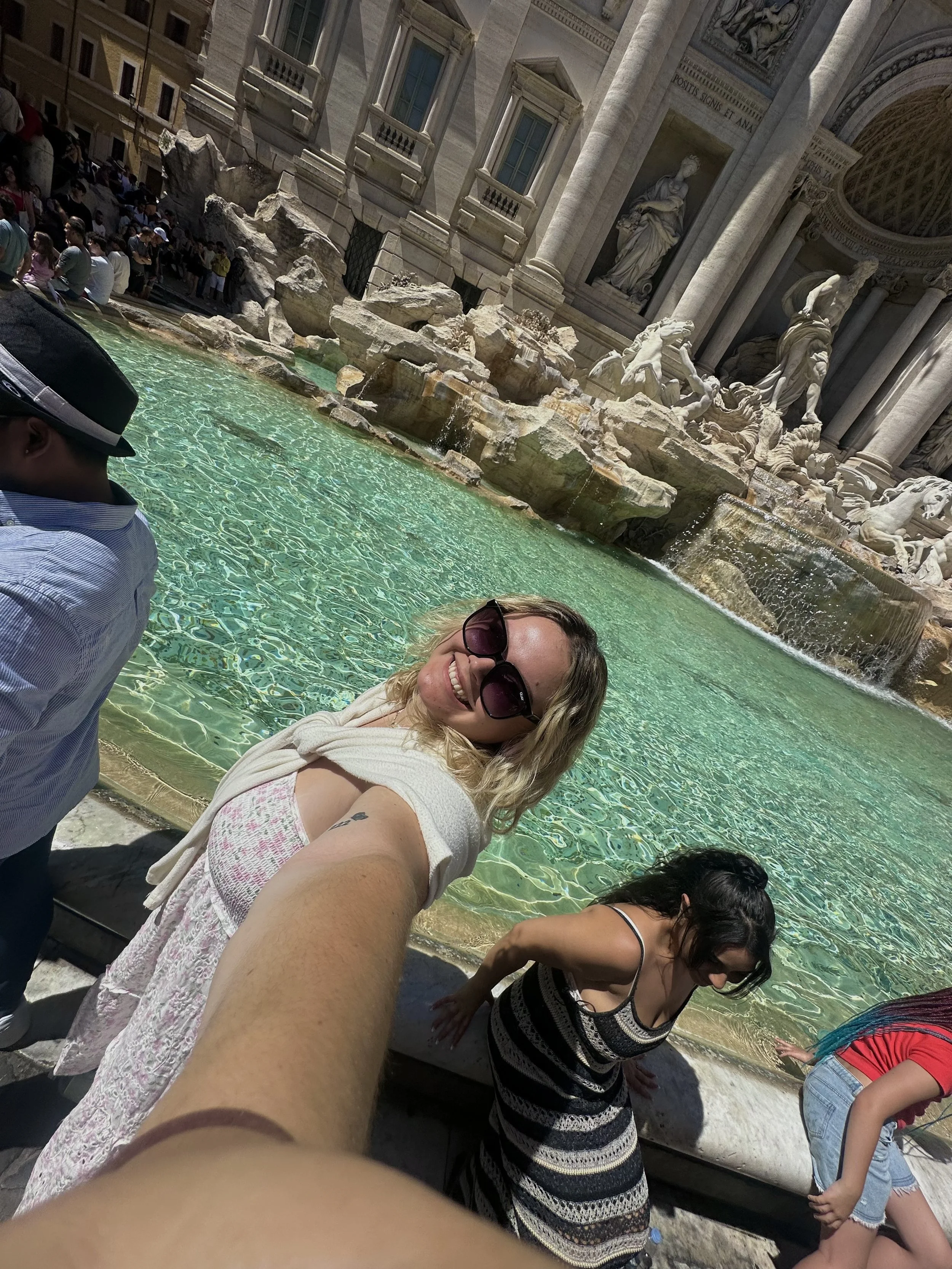 People standing and sitting around the Trevi Fountain in Rome, Italy, with classical statues and elaborate architecture in the background.