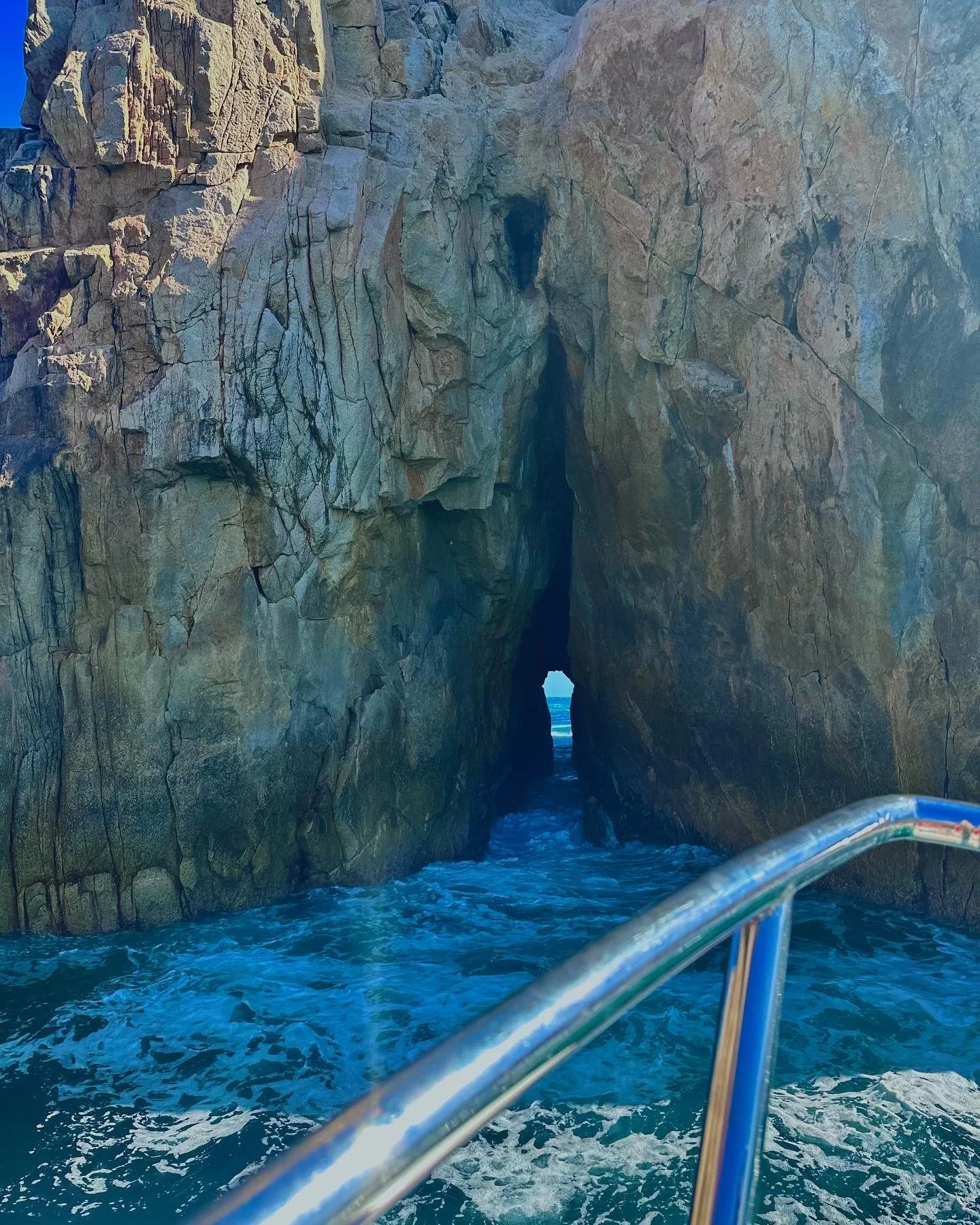 Photo taken from a boat showing a natural sea arch formation in a rocky cliff with waves crashing at the base.