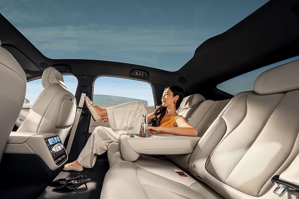 A woman relaxing in the backseat of a luxury vehicle with beige leather seats, a panoramic sunroof, and a view of the sky and clouds outside.