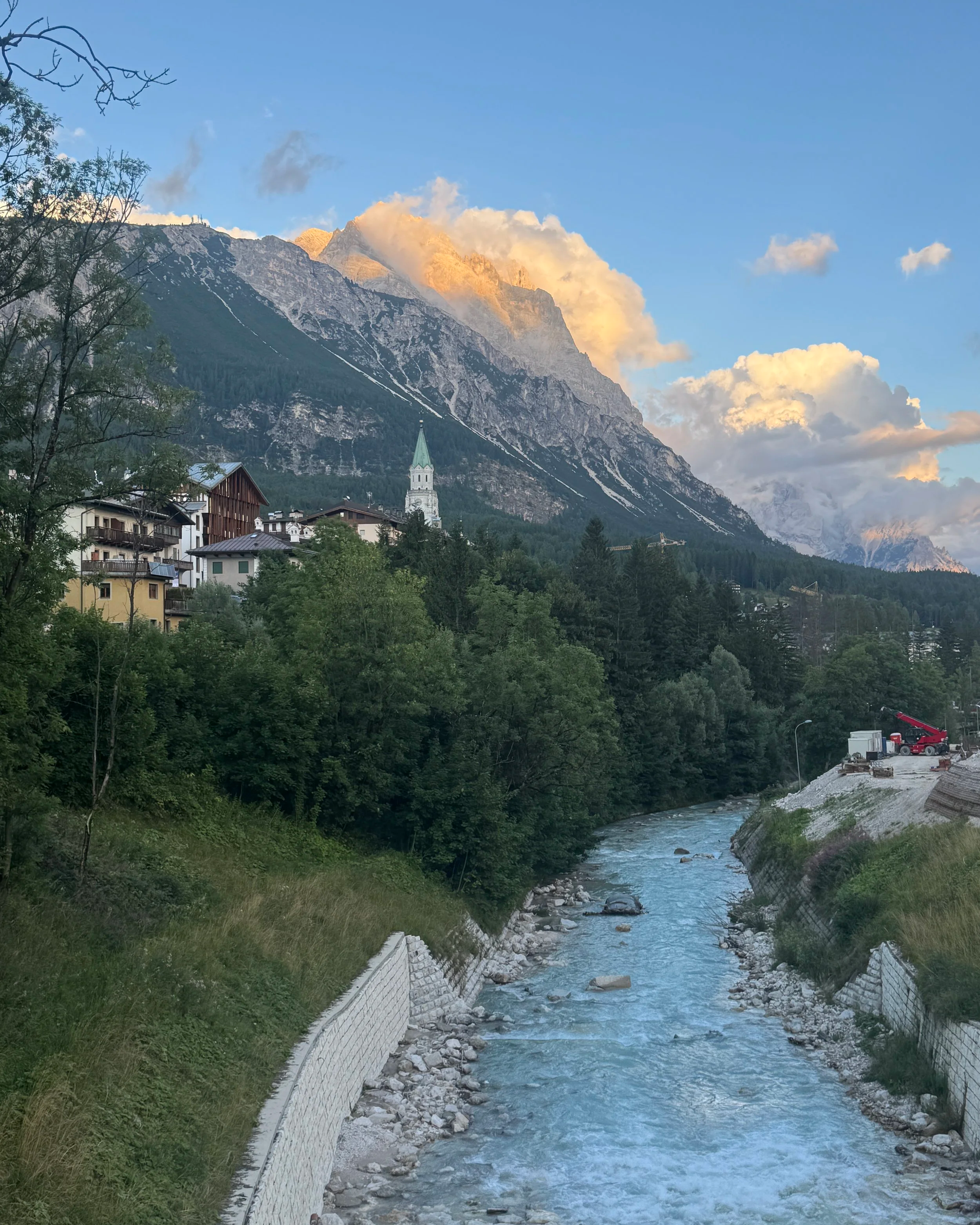 A mountain river flowing through a green valley with a small village at the base and a church steeple. Tall mountains with rocky slopes and patches of snow are in the background, with clouds and sunlight illuminating the peaks.