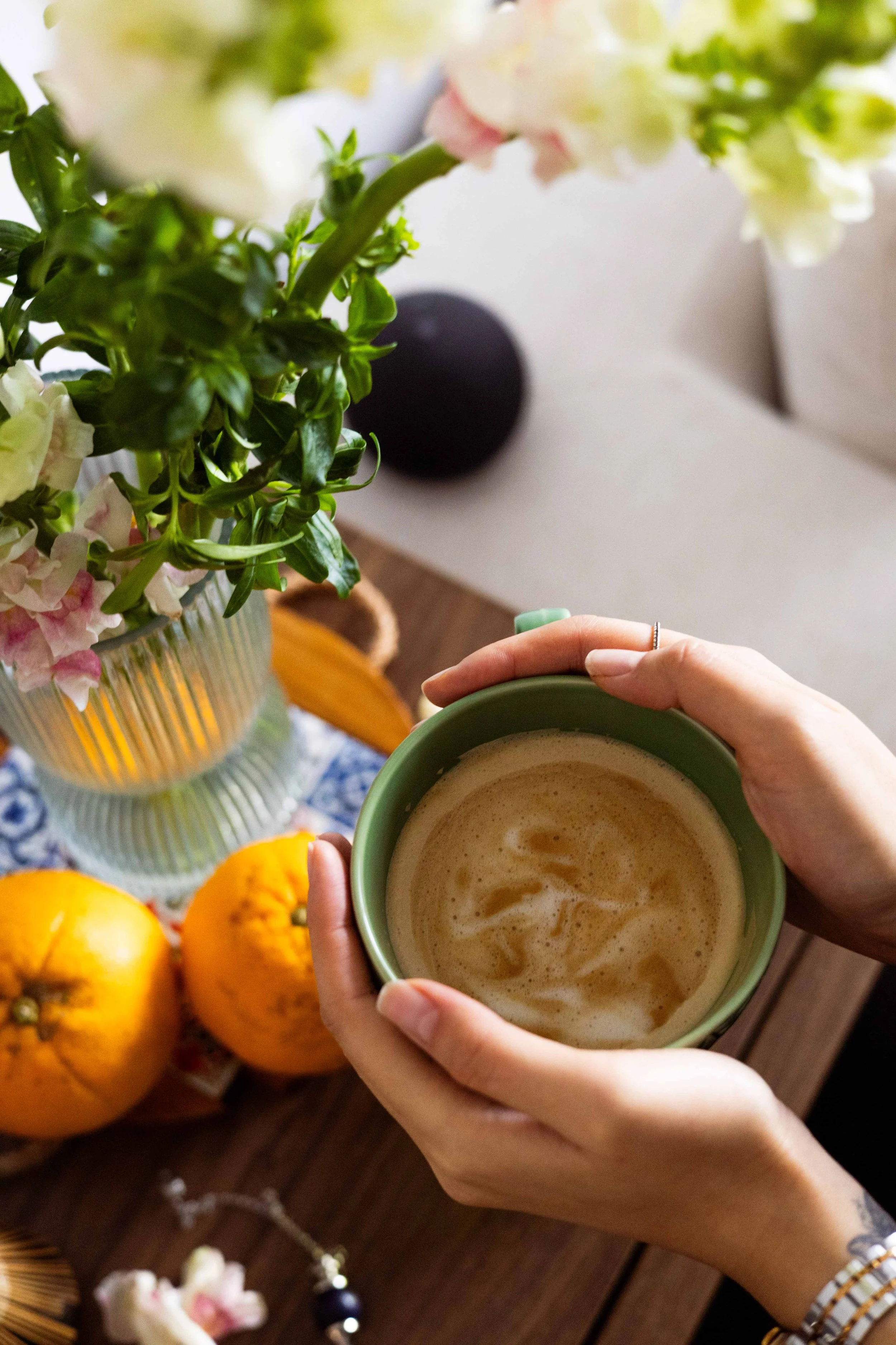 Hands holding a foam-topped coffee, with oranges and flowers on a wooden table.