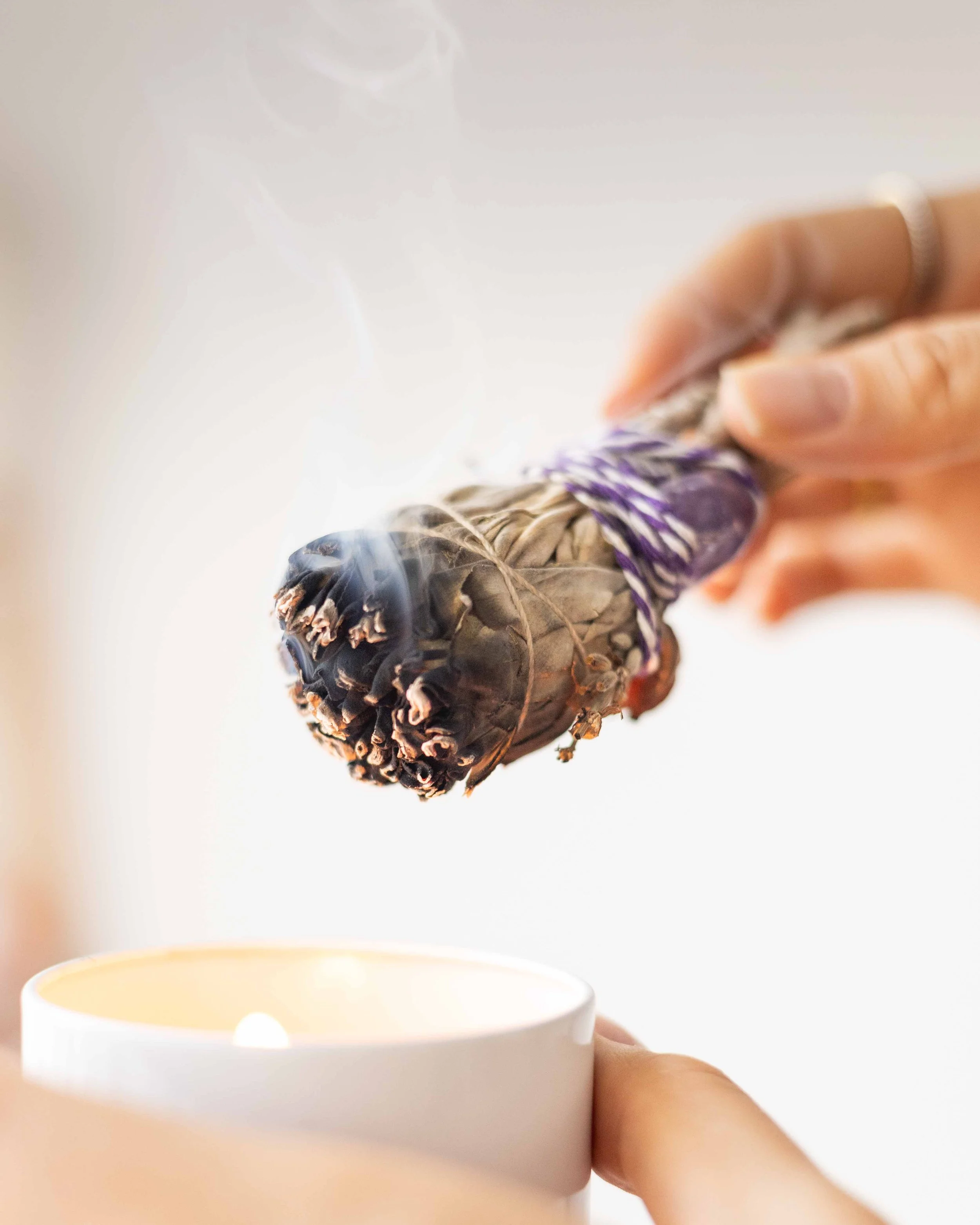 Hands lighting a bundle of dried herbs over a candle flame.
