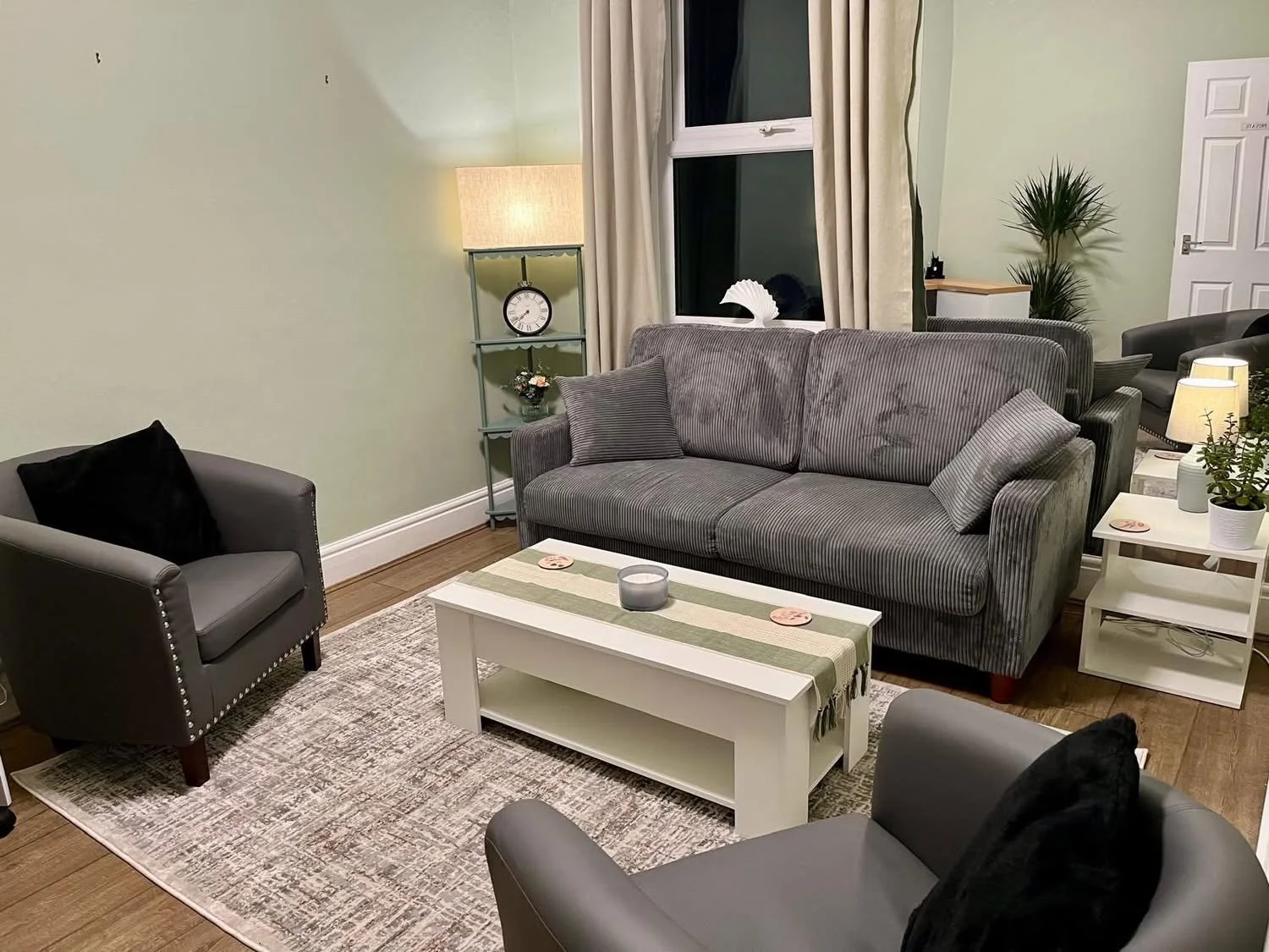 Living room with gray striped sofa, black armchair, coffee table, side table with plants, bookshelf, lamp, and window with curtains.