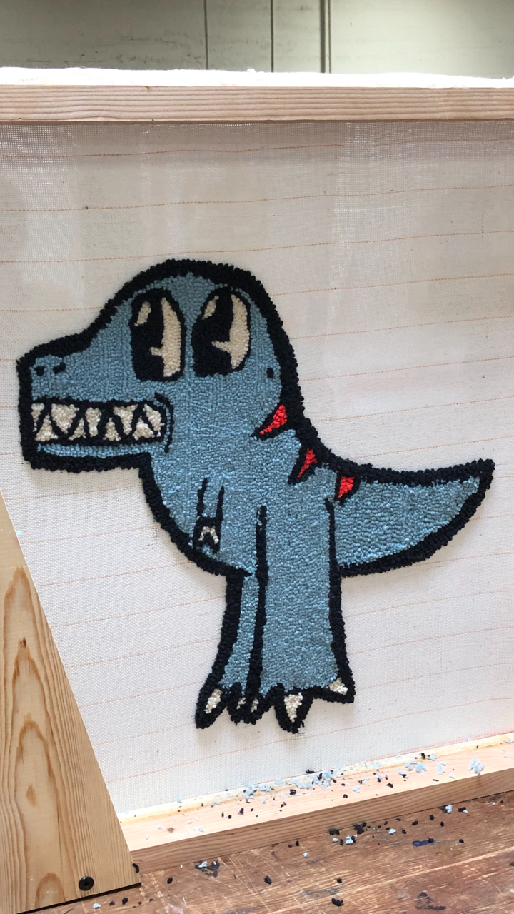 A  tufted rug on its whie canvas showing a blue, cartoon dinosaur with sharp teeth and black outline.
