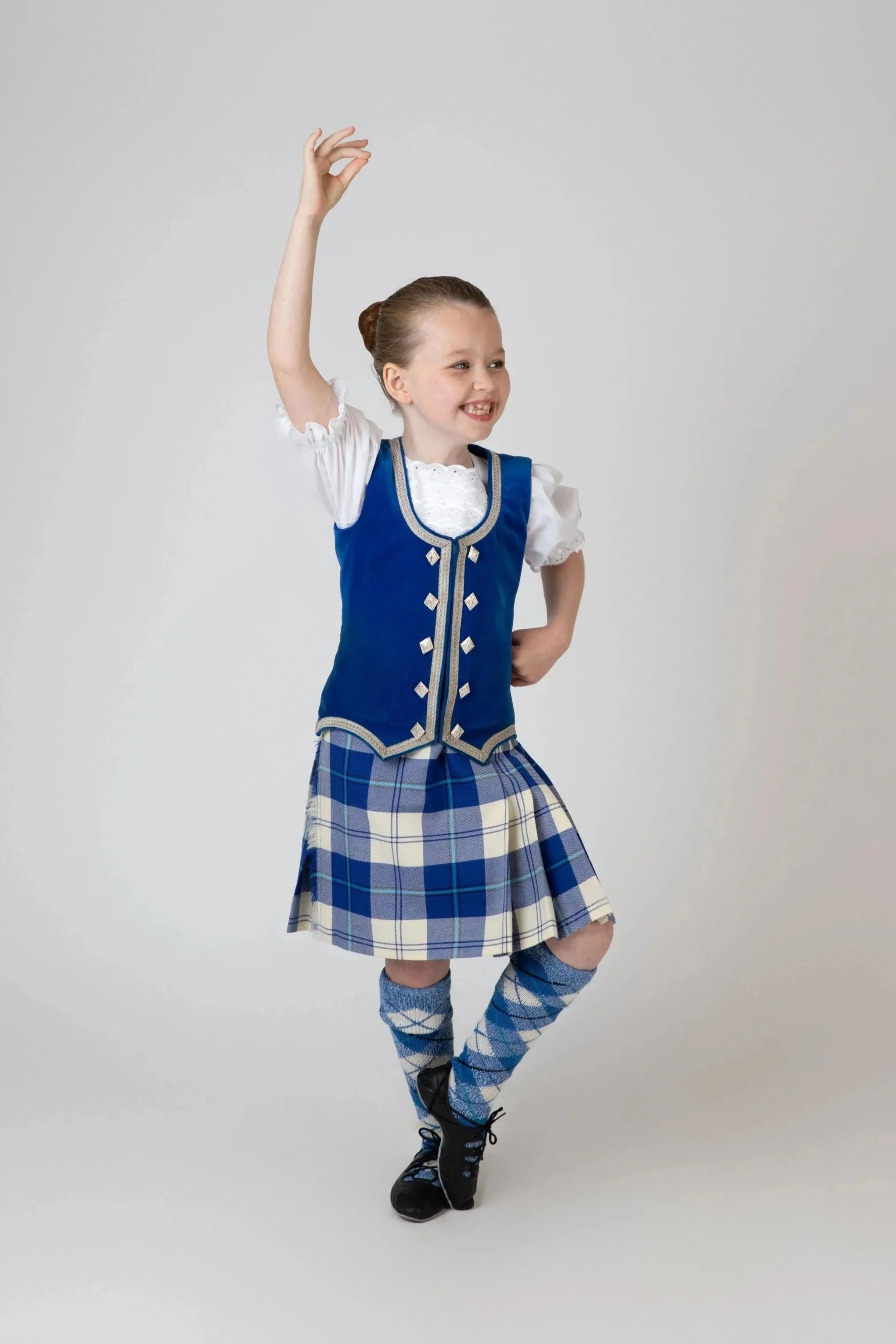 A young girl with red hair tied back, wearing a traditional Scottish outfit including a blue vest with silver buttons, a plaid skirt, knee-high argyle socks, and black boots, is standing against a plain white background. She is smiling and raising her right arm with her hand positioned as if doing a dance move.