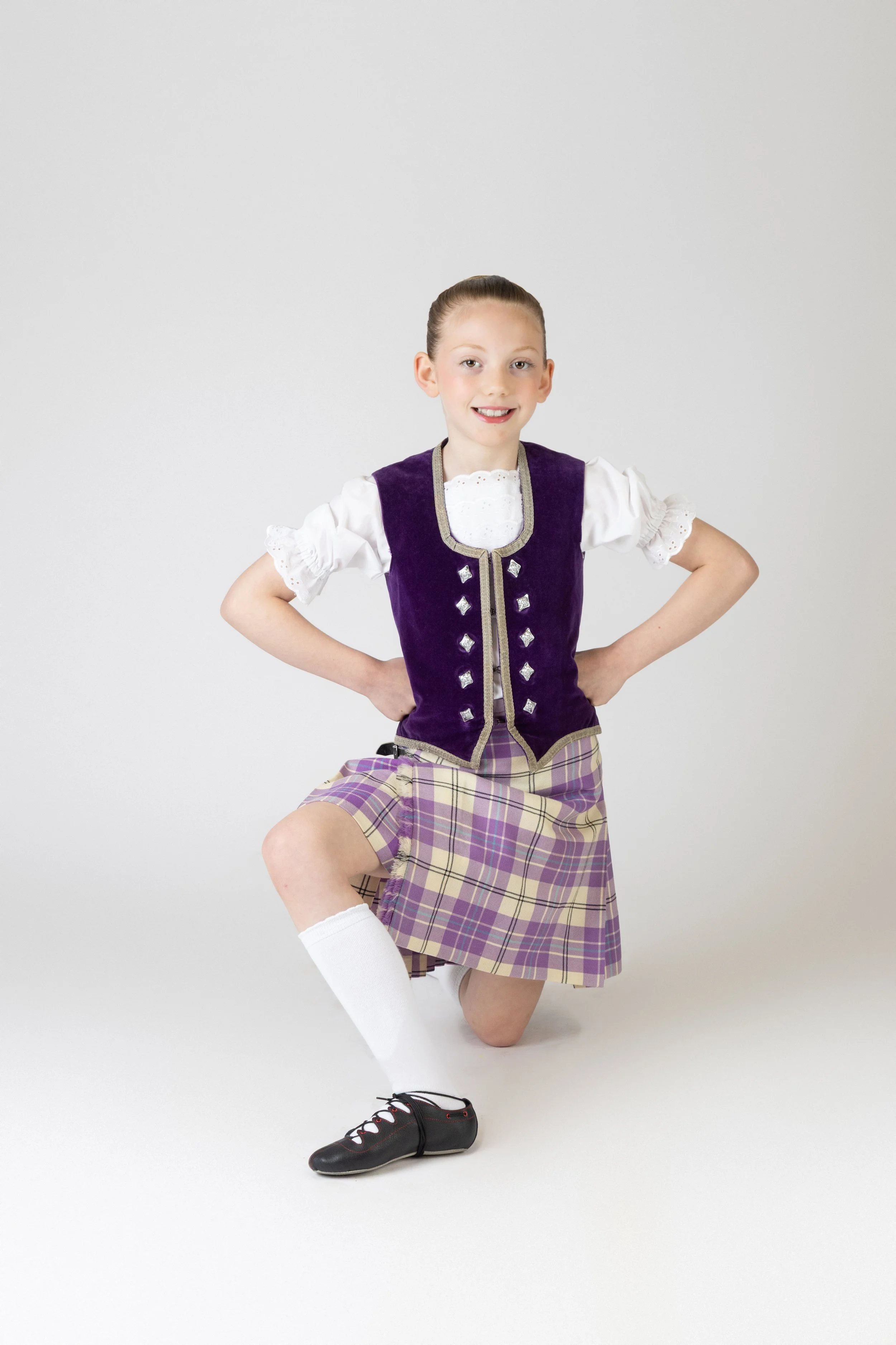 A girl with her hair in a bun, wearing a purple vest with silver trimming and buttons, a white blouse with puffed sleeves, a plaid skirt, white socks, and black shoes, posing with one knee on the ground and her hands on her hips against a plain white background.