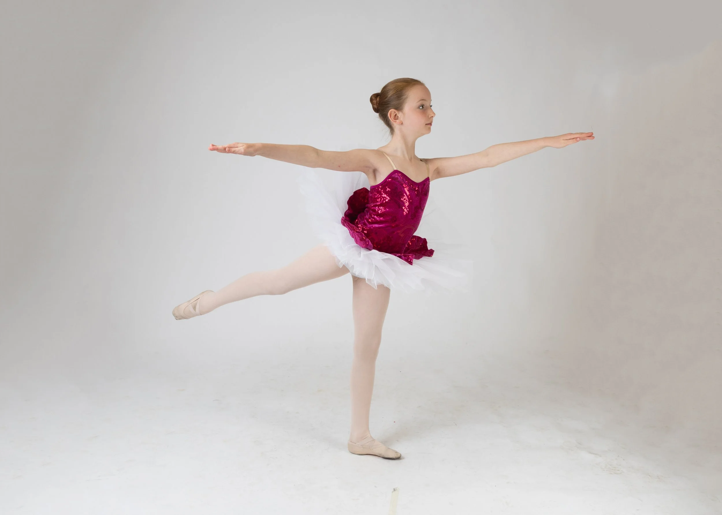 A young girl with red hair tied in a bun, wearing a pink sequined leotard and a white tutu, is performing an arabesque ballet pose on pointe shoes against a plain gray background.