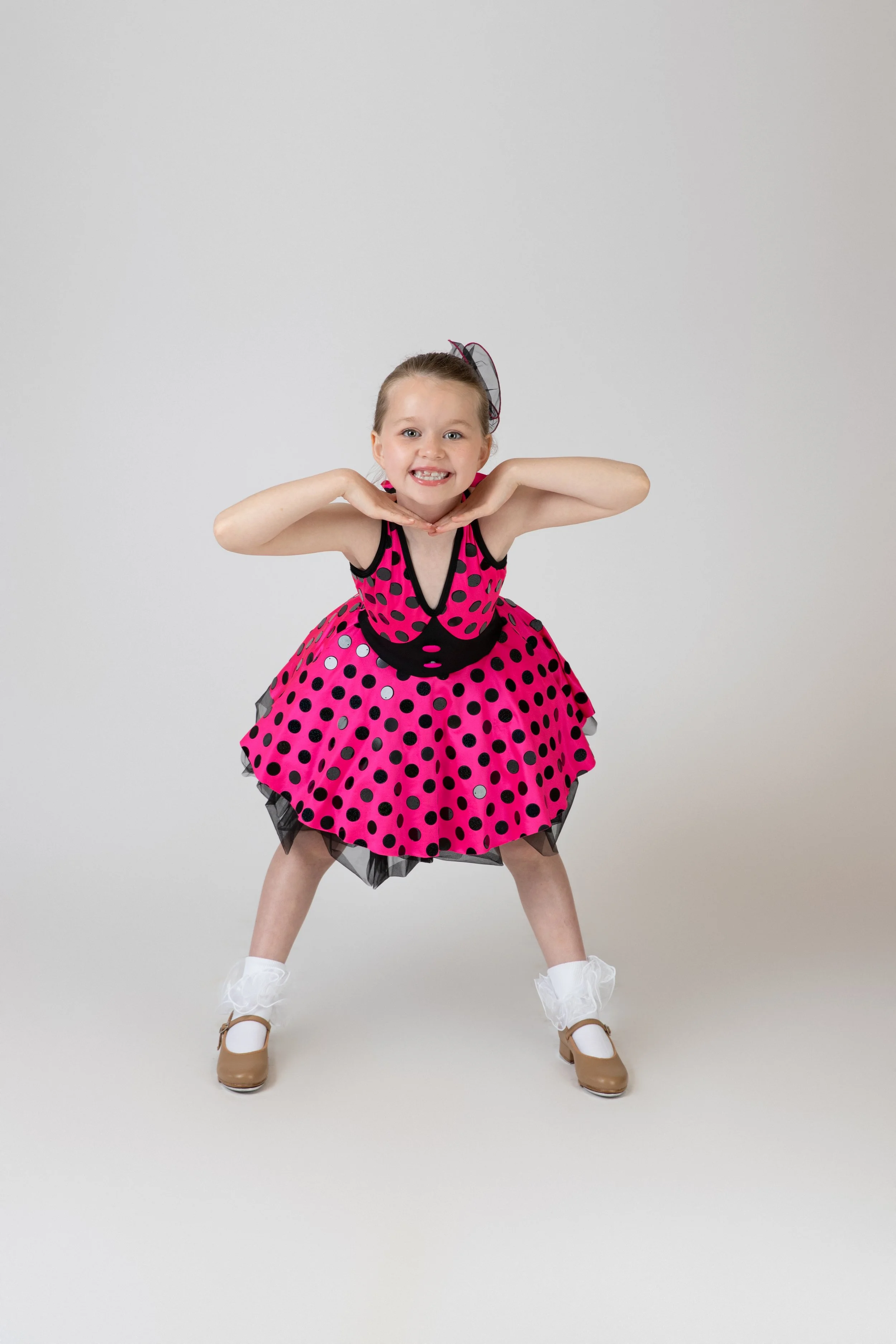 Young girl in a pink and black polka dot dress with a tulle skirt, wearing white socks and tan shoes, standing in a dance pose with hands under her chin and smiling against a plain gray background.