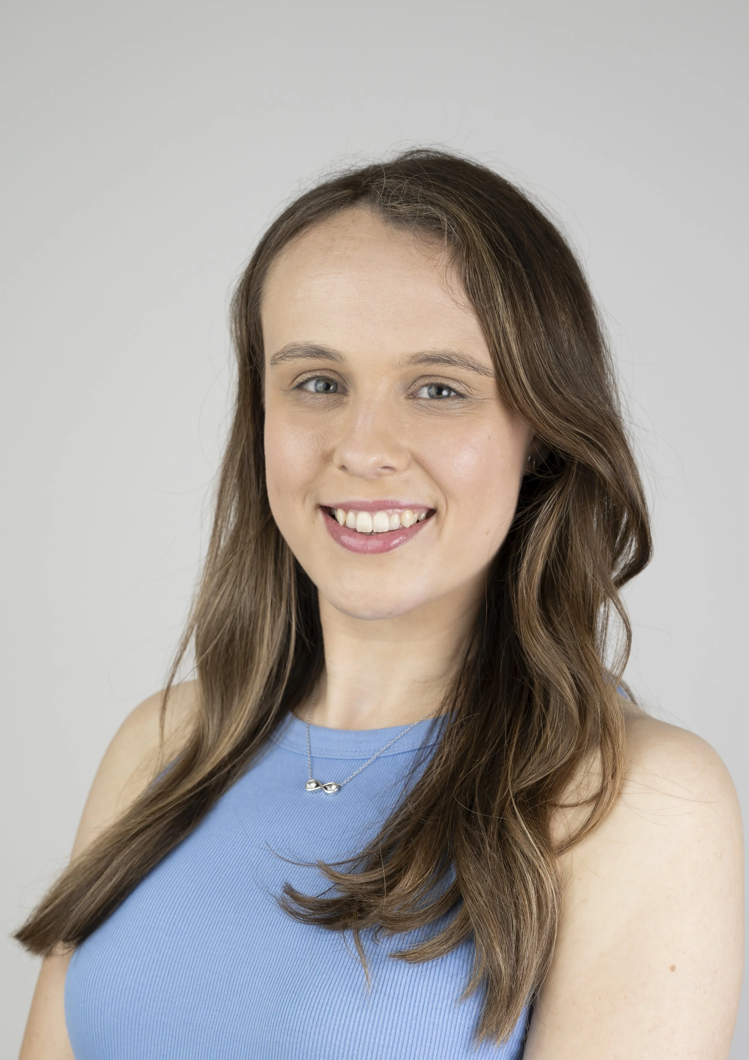 A young woman with long, wavy brown hair and blue eyes, smiling, wearing a sleeveless light blue top and a silver infinity necklace against a plain gray background.