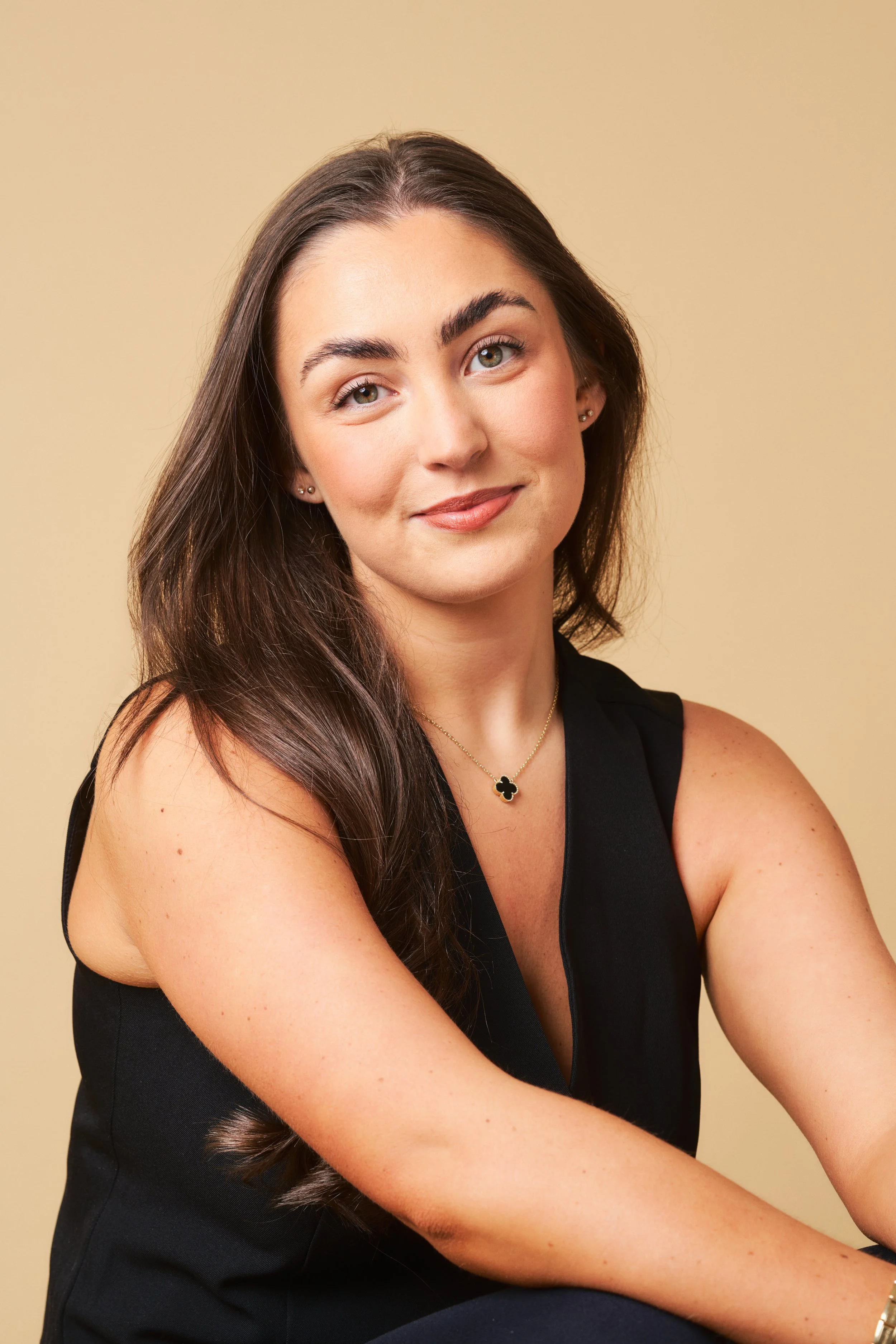A young woman with long dark hair and light skin, wearing a black sleeveless top, gold necklace with a four-leaf clover pendant, and small earrings, smiling with her arms crossed against a beige background.