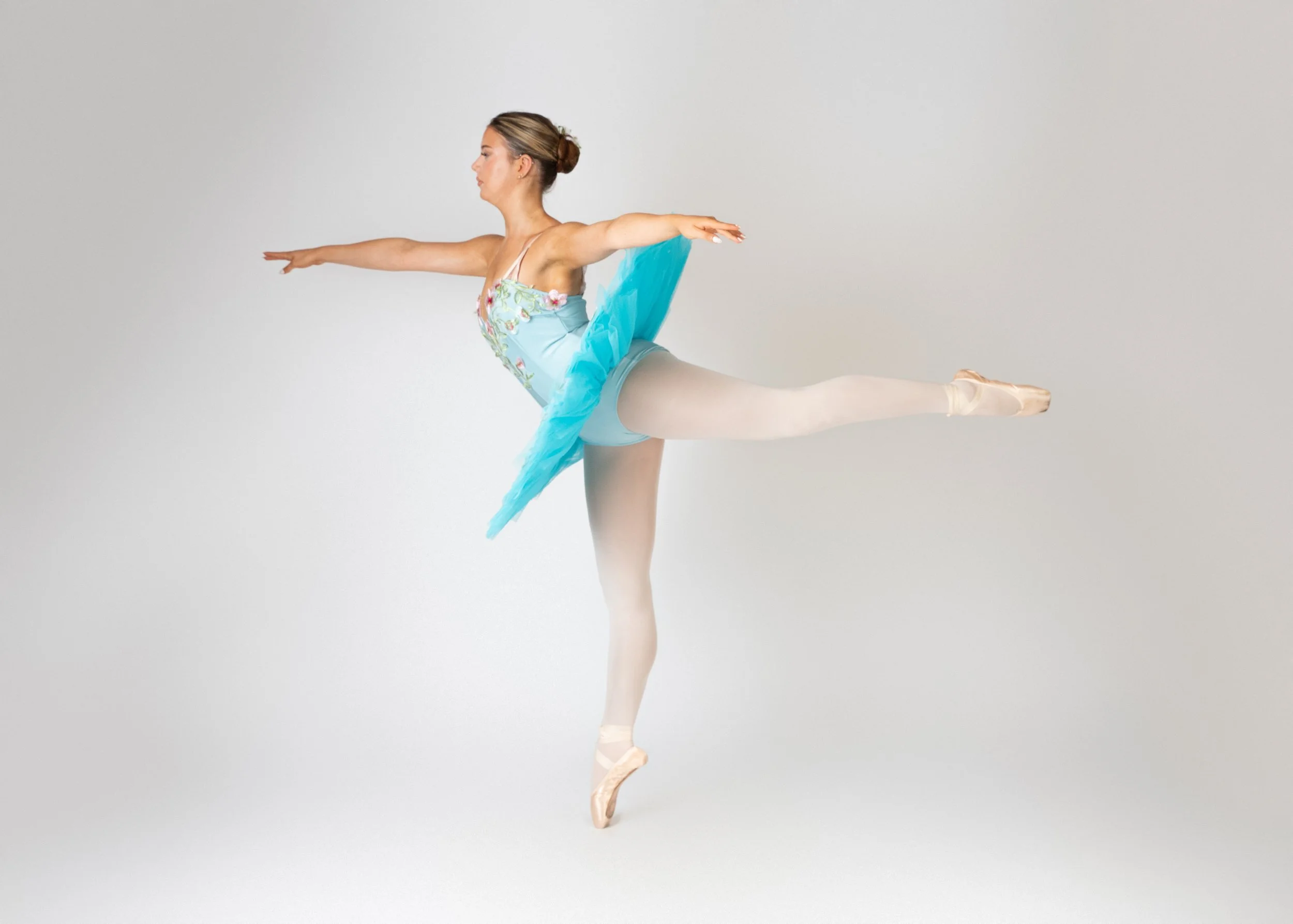 A ballet dancer performing an arabesque pose, standing on one leg with the other leg extended behind her and arms stretched forward, against a plain white background.