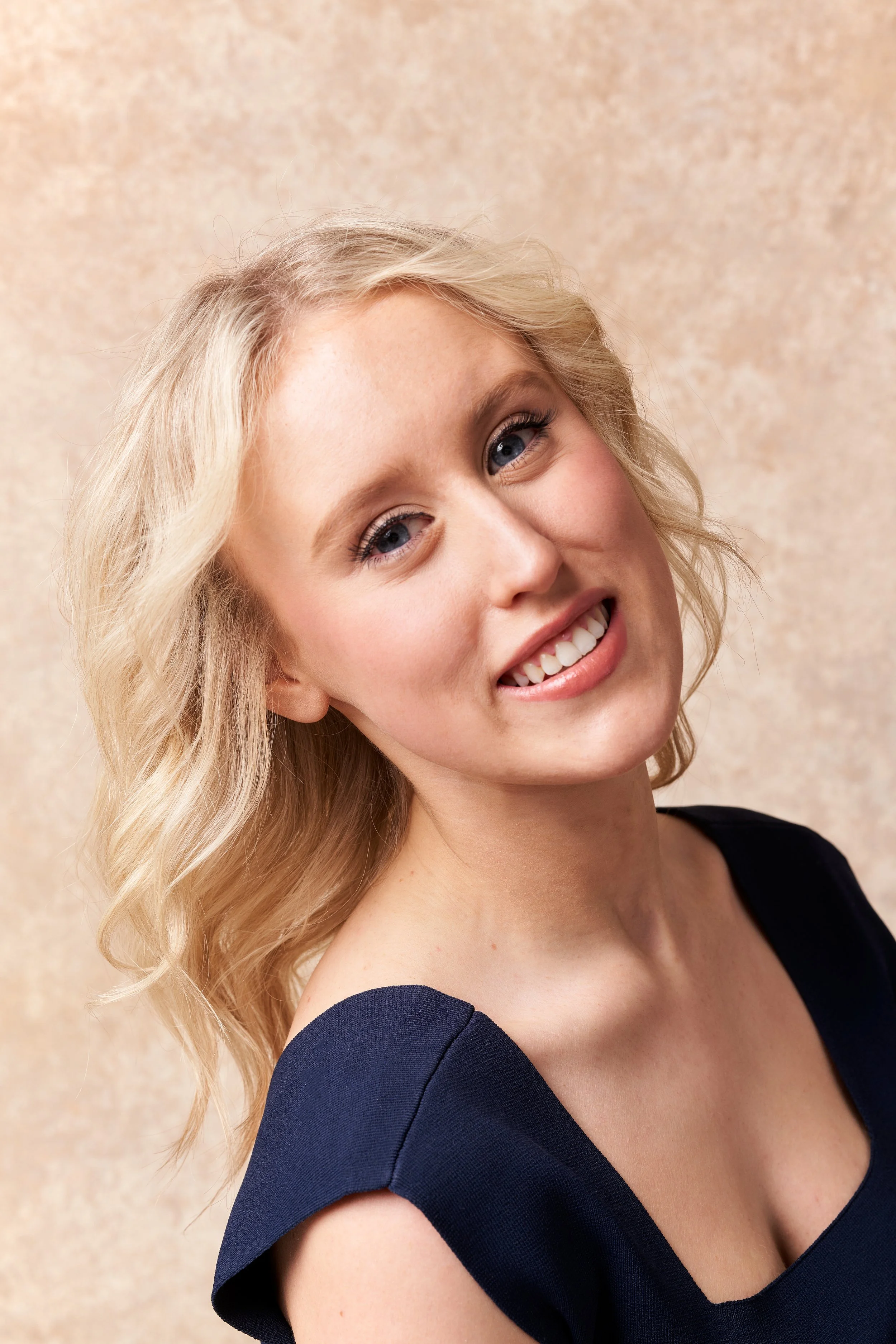 Close-up of a woman with blonde hair smiling and wearing a black dress with ruffled sleeves, standing against a beige textured background.