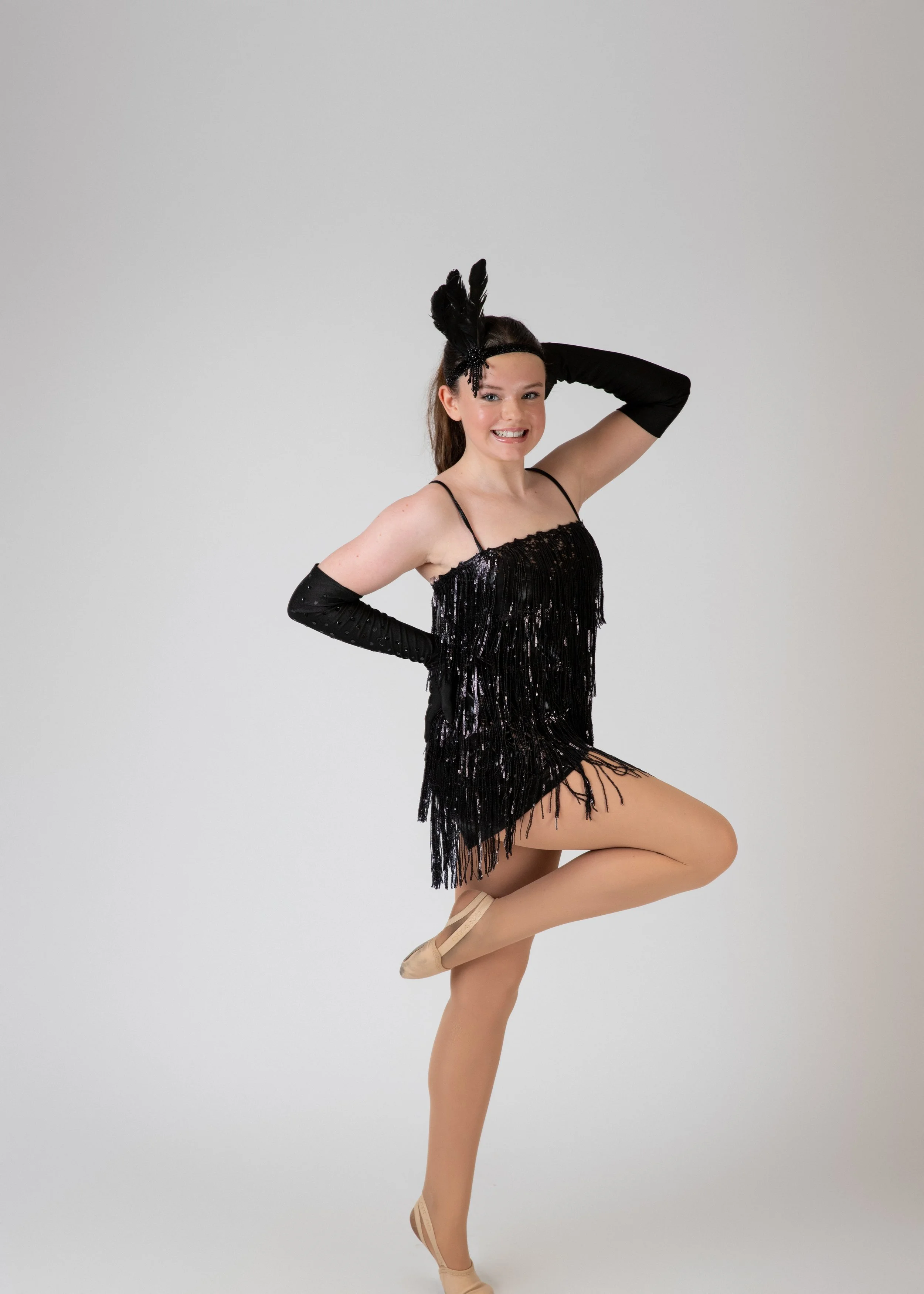 A young woman in a black fringe dress, wearing tan dance shoes and black gloves, poses on her toes in a ballet stance with one arm raised and the other hand on her head, against a plain white background.
