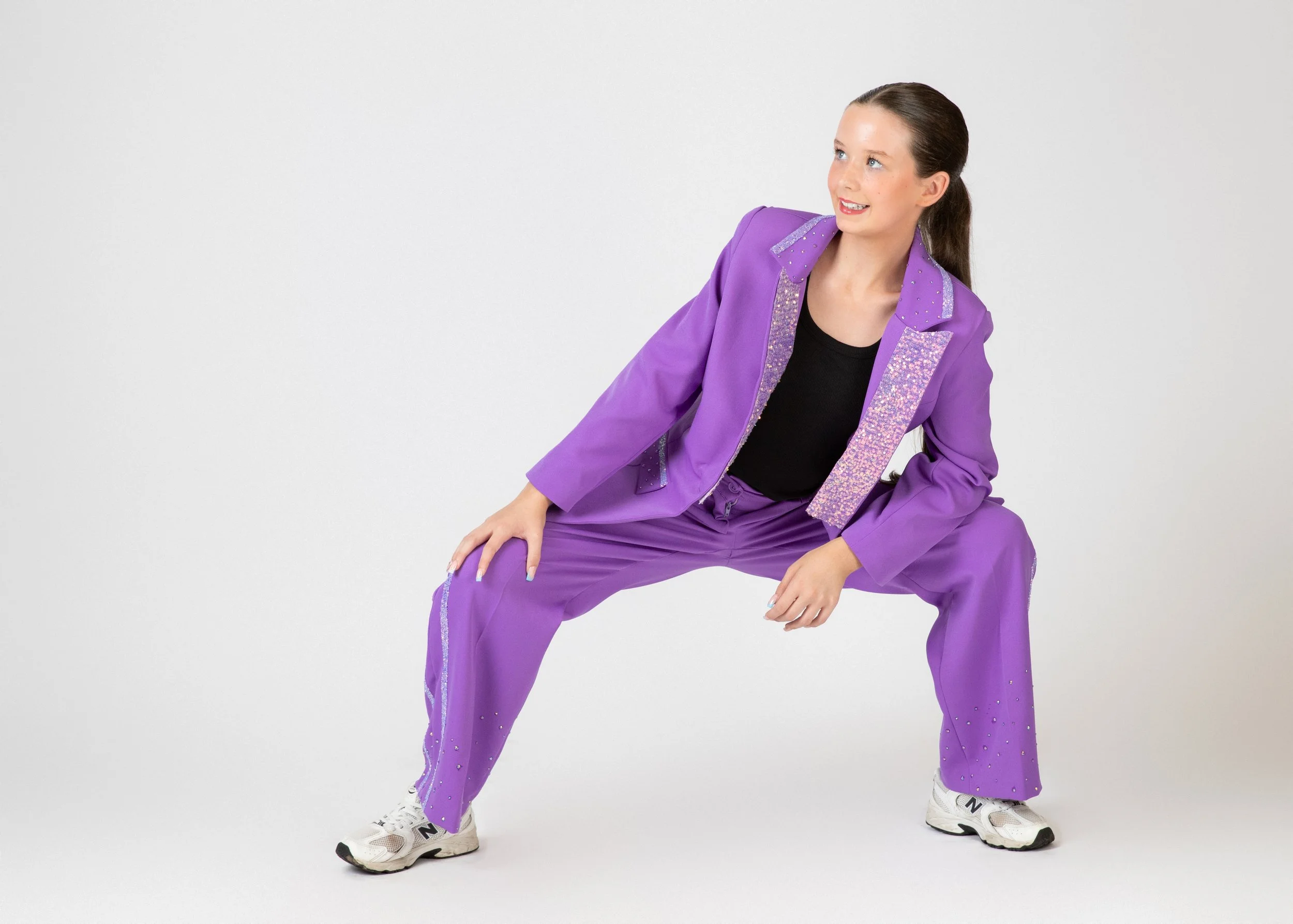 A woman in a purple sequined jacket and matching pants poses with a wide stance, wearing athletic shoes, against a plain white background.