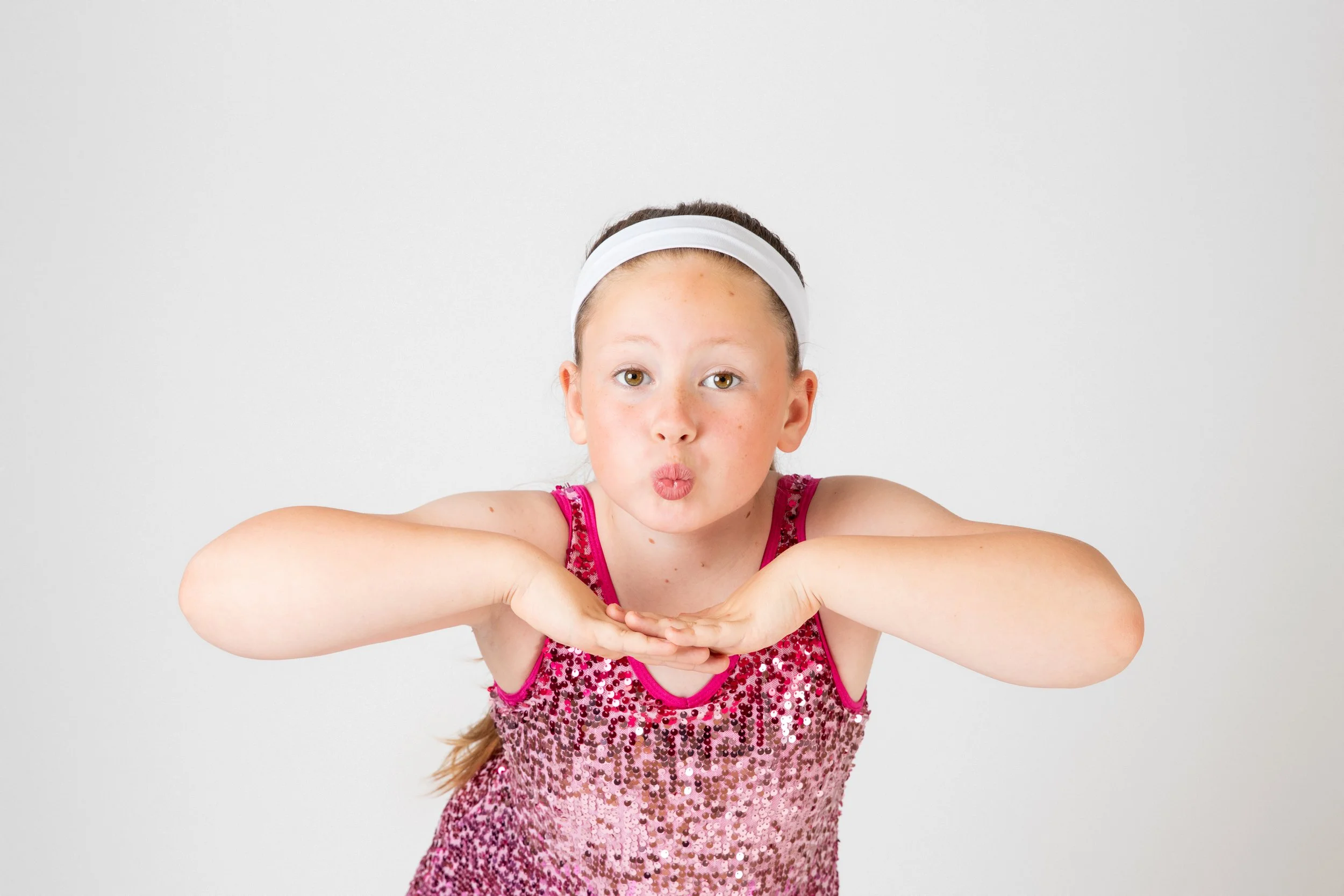 A young girl wearing a pink sequin dress and a white headband, making a playful pose with her arms extended forward and lips puckered.