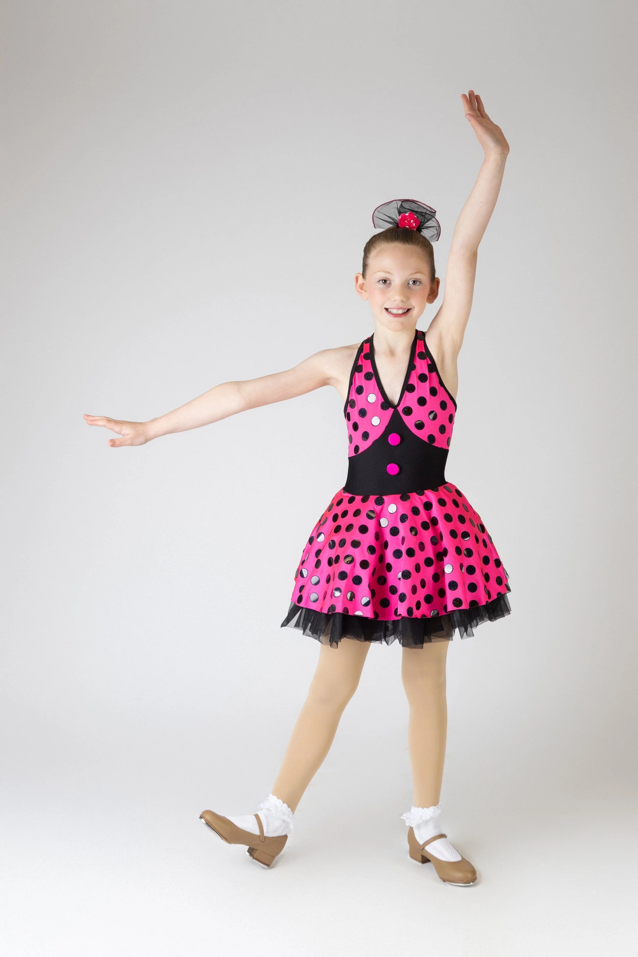 Girl wearing a pink and black polka dot floral dress, hair in a bun with a black netted bow, balancing on one foot with arms raised, wearing tan tap shoes, beige tights, and white socks.