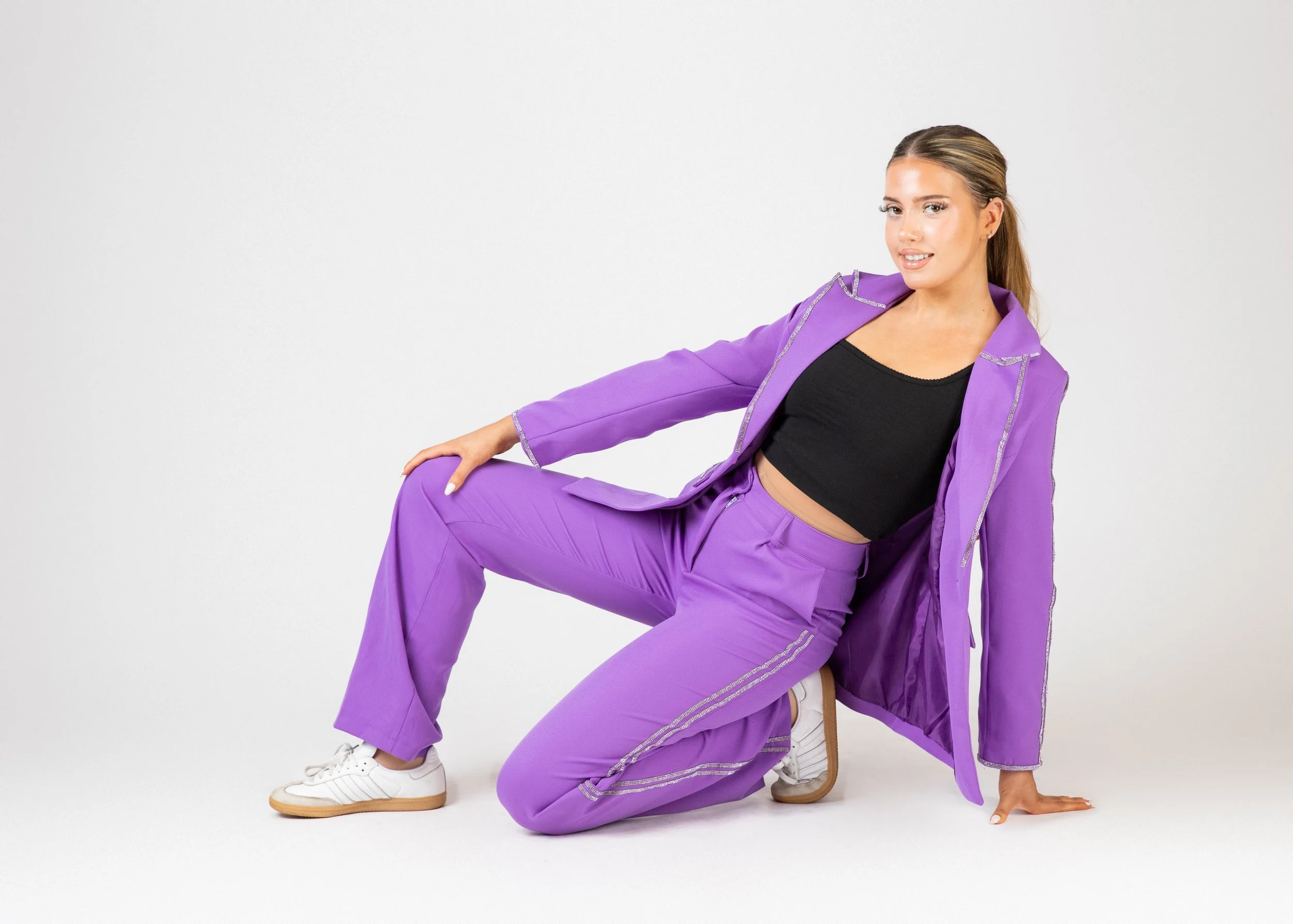 Woman in purple suit and black crop top poses on the floor against a plain white background.