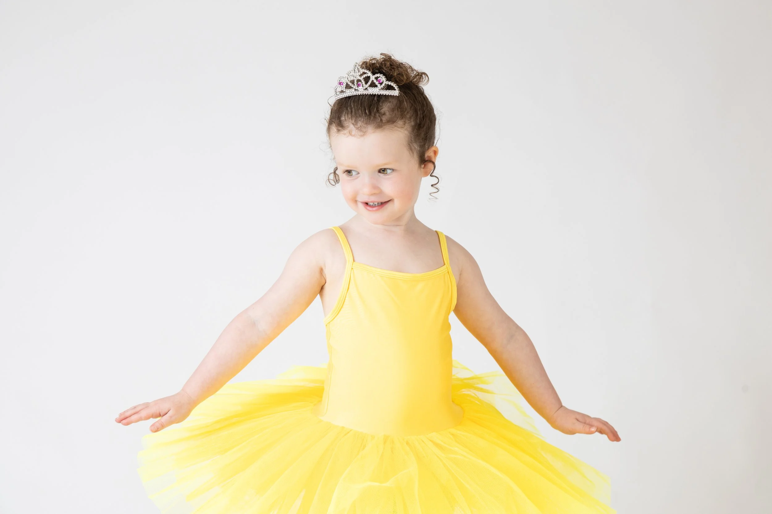 A young girl dressed as a princess in a yellow dress with a tiara, sitting against a plain white background.
