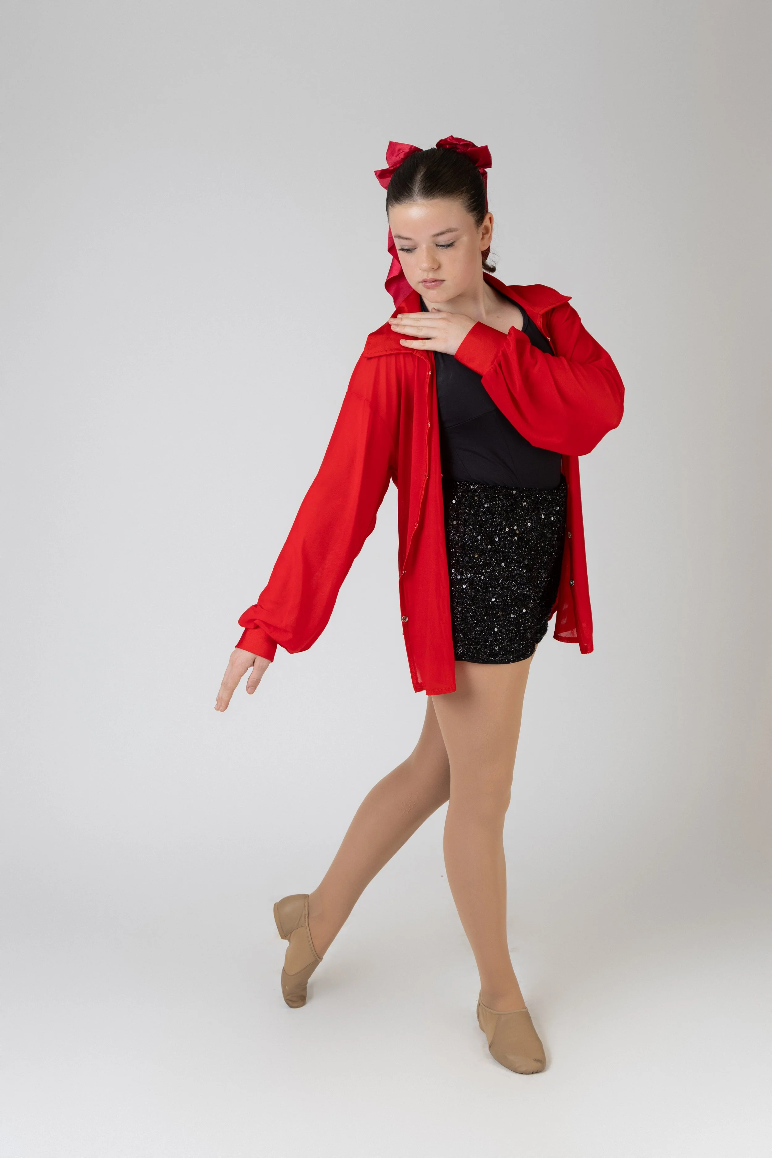 Young female ballet dancer in beige dance shoes, black leotard, red jacket, and red bow in her hair, striking a balletic pose against a plain white background.