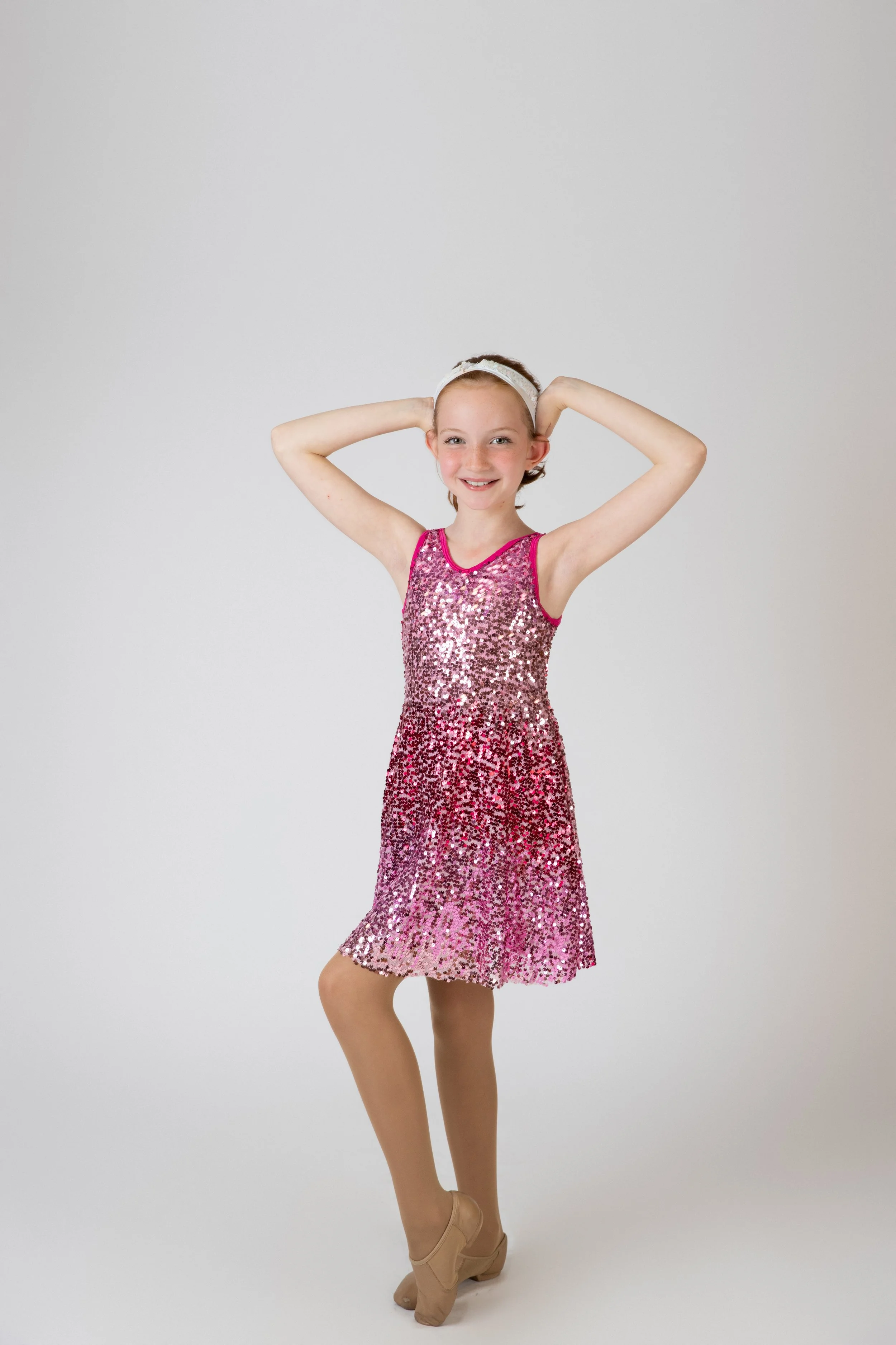 A young girl in a pink, sequined dress with a gradient from light to dark pink, standing in a dance pose with her hands behind her head against a plain gray background.