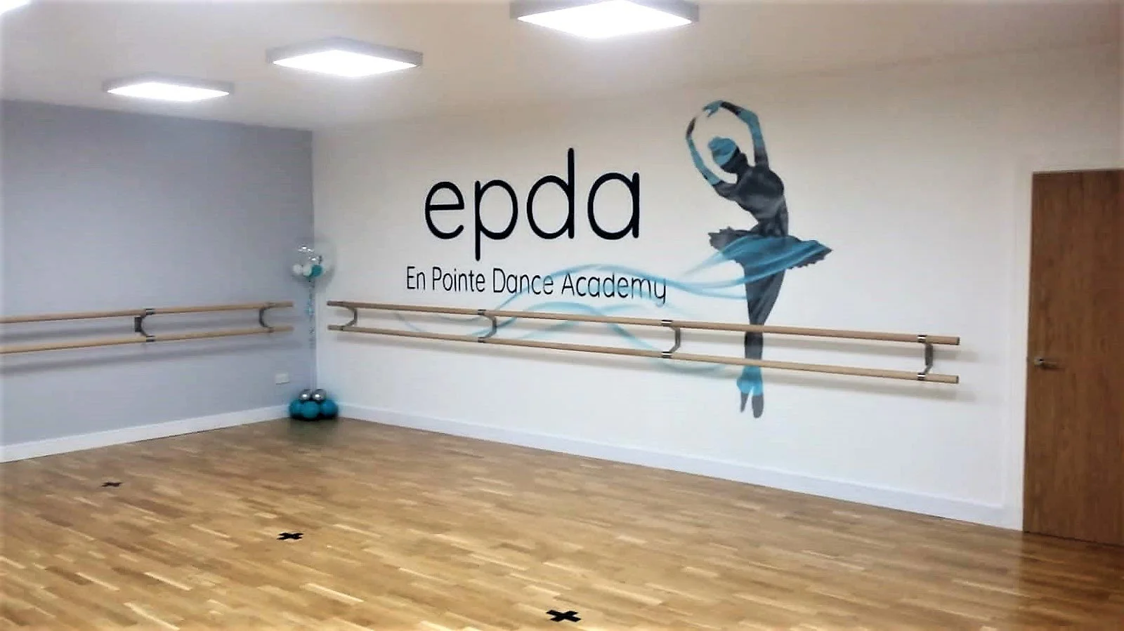 Dance studio with wooden floor, ballet barre, and wall art featuring a ballerina and the text 'epda En Pointe Dance Academy'.