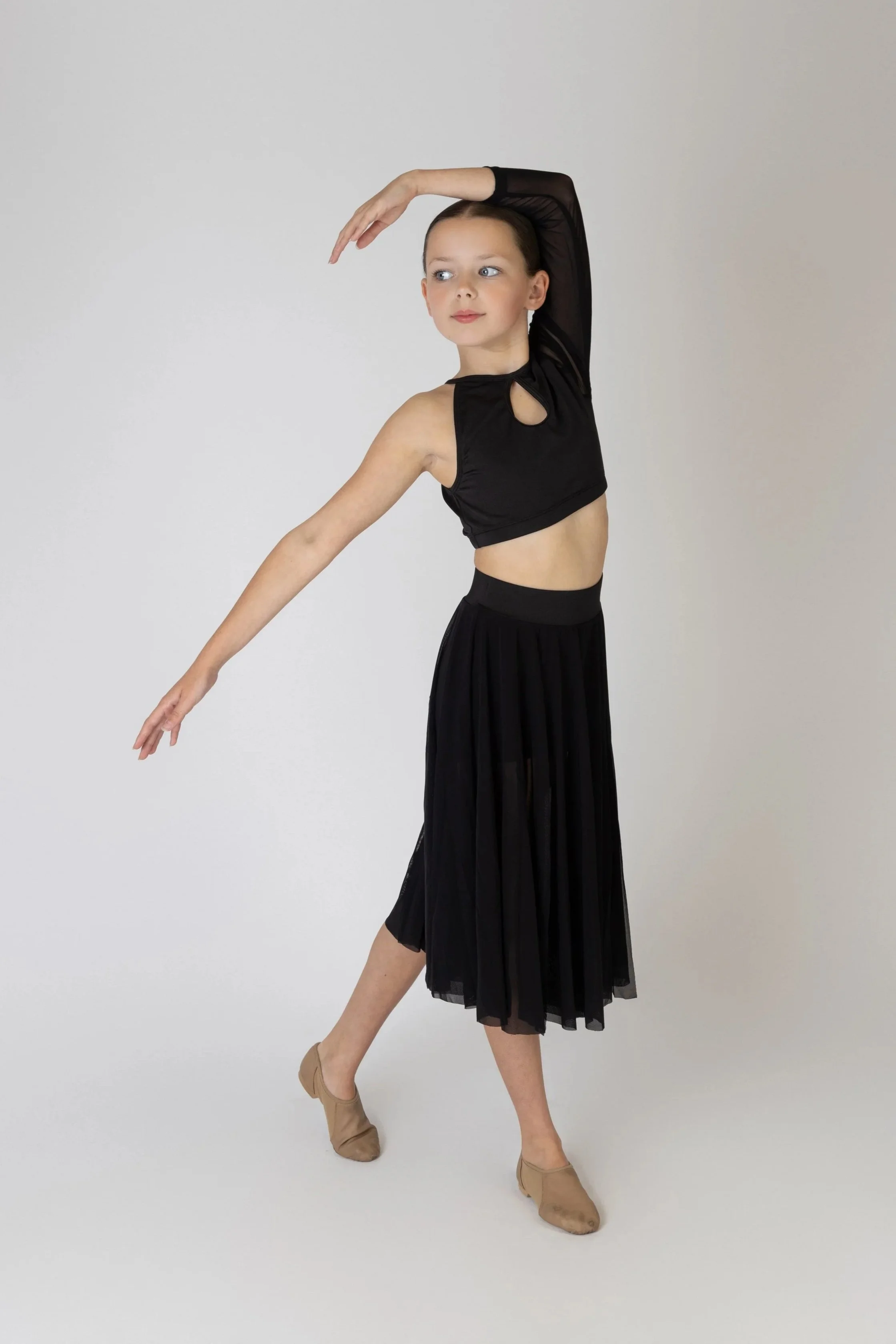 A young girl dancing, wearing a black cutout top with long sheer sleeves, a long black skirt, beige dance shoes, and balancing on one leg with arms extended, against a plain white backdrop.
