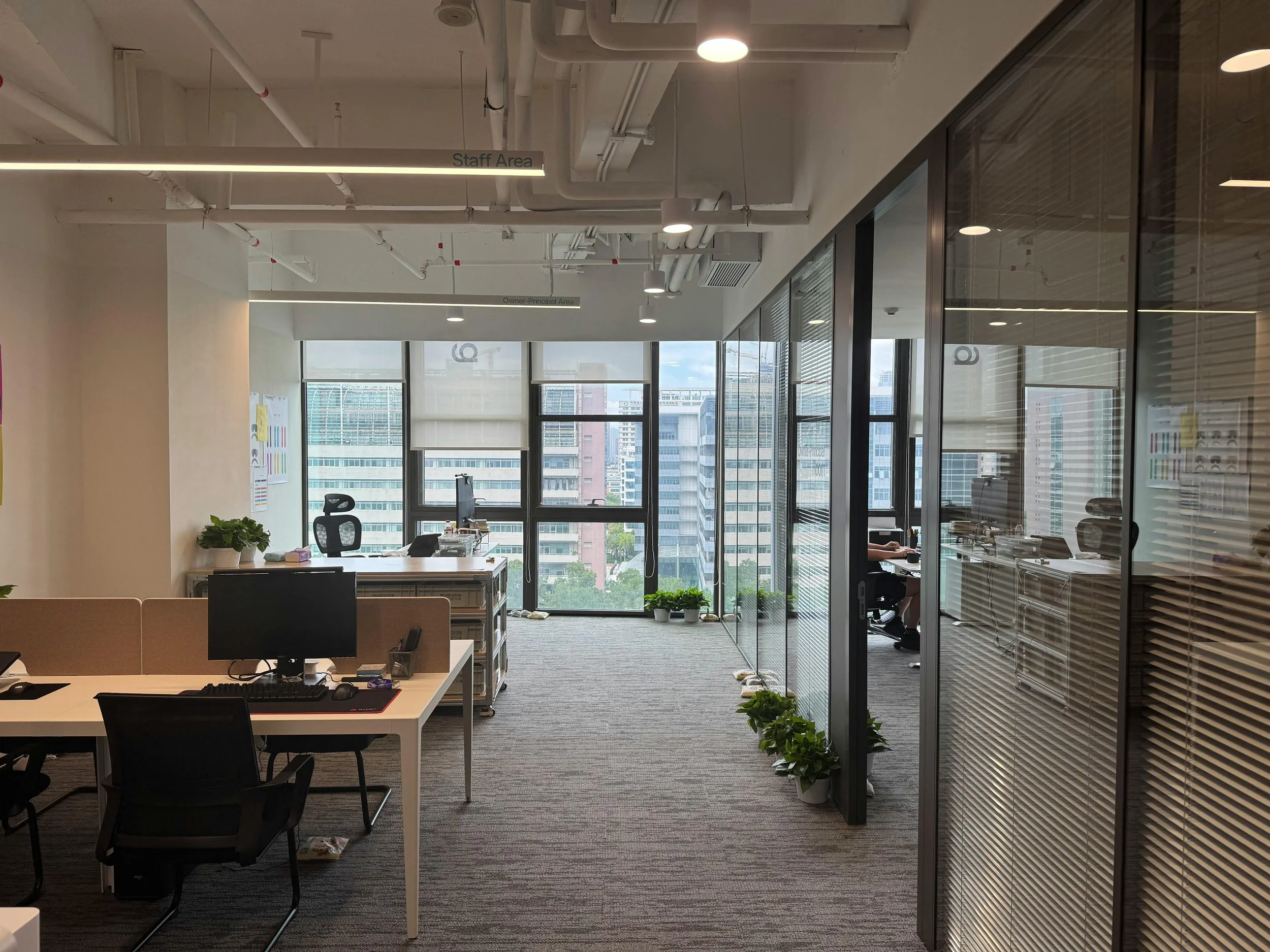 An office space with white walls and a large window showing a city skyline. Multiple work desks with computers and office chairs are visible, along with potted plants on the floor. Overhead lighting and ceiling pipes are also present.