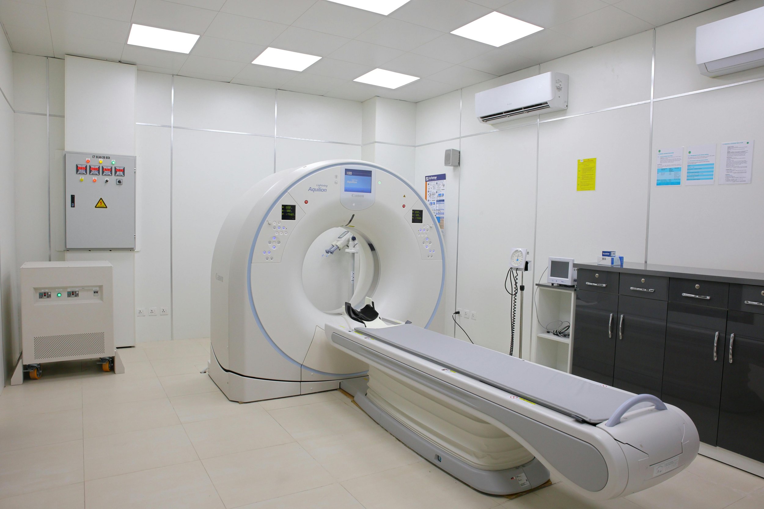Medical imaging room with a large CT scan machine, control panels, and medical equipment.