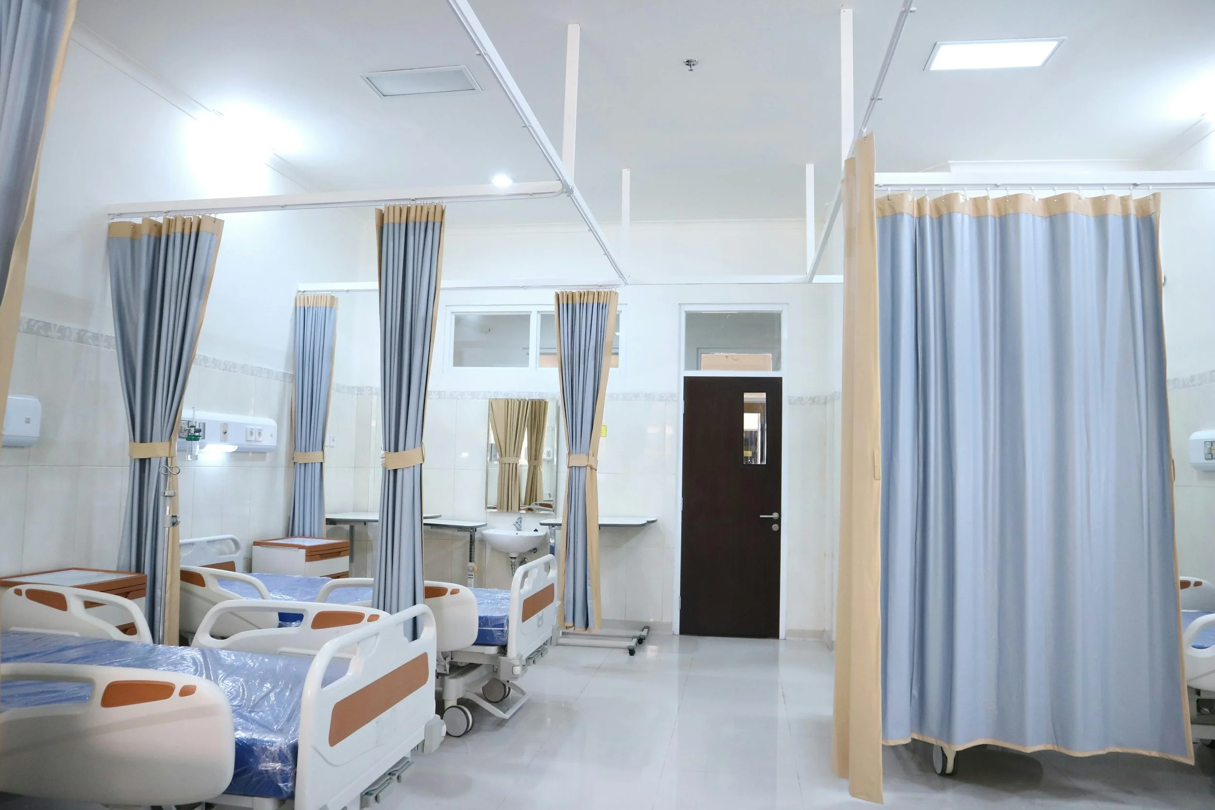 Hospital patient room with several empty beds separated by blue and beige privacy curtains, a sink, mirror, and door in the background.