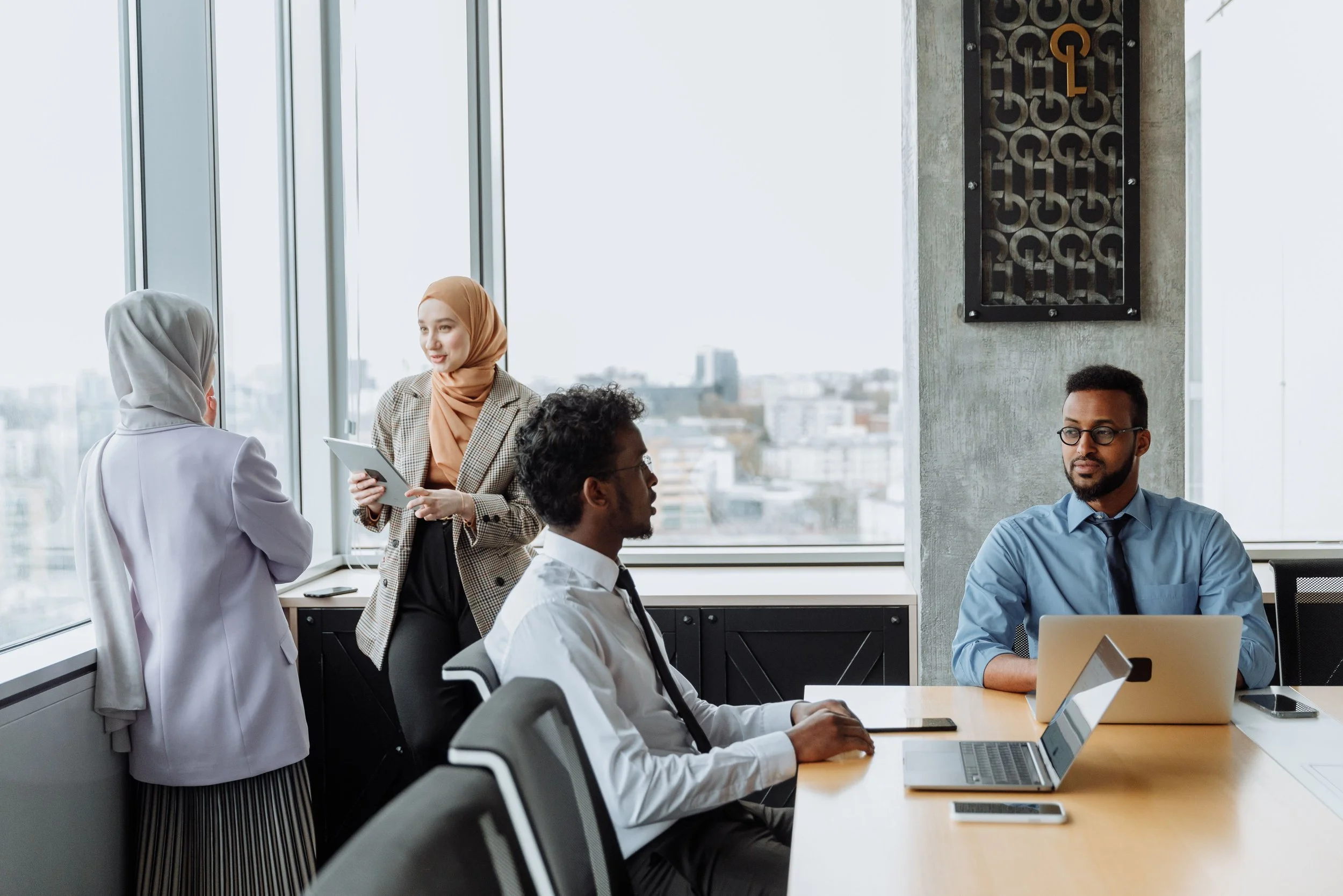 A diverse group of professionals in a modern office with large windows, engaging in conversation and working on laptops.