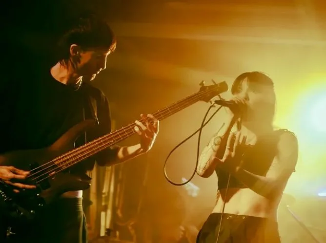 A male musician playing bass guitar and a female singer performing with a microphone on stage, illuminated by warm yellow stage lighting.