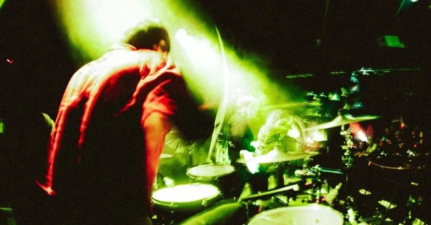 A drummer performing on stage with green and yellow lighting, surrounded by a live crowd.