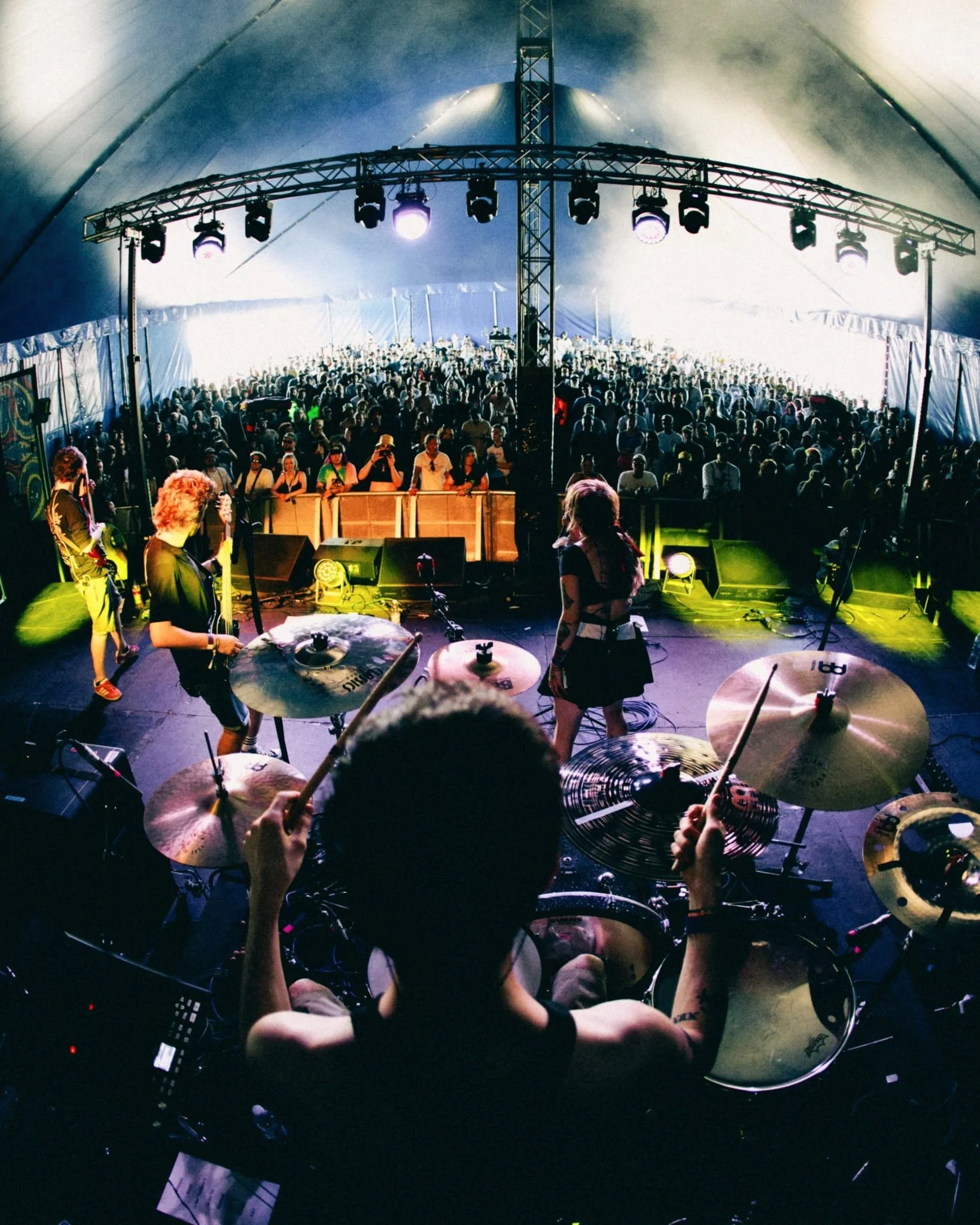 A band performing on stage with drums, guitar, and multiple musicians, viewed from behind the drummer in a large tent with an audience in front.