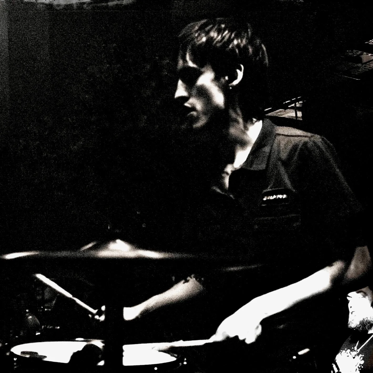 Black and white photo of a person with a short haircut, wearing a dark shirt, working on a DJ turntable in a dimly lit environment.