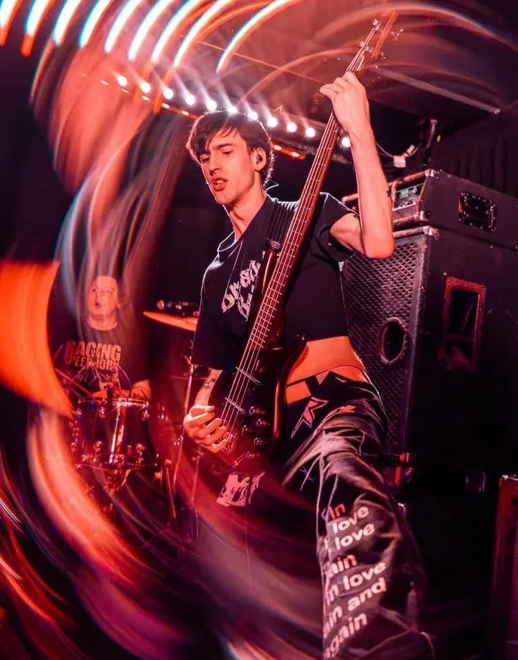 A young male musician playing a bass guitar on stage with colorful light streaks around him, in a vibrant concert setting.