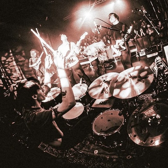Band performing live on stage with musicians playing guitars and drums, and an audience watching in the background.