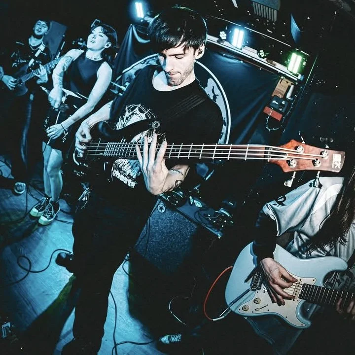 Musicians performing live on stage at a club, with one playing an electric bass guitar and others with guitars, under blue and green stage lighting.