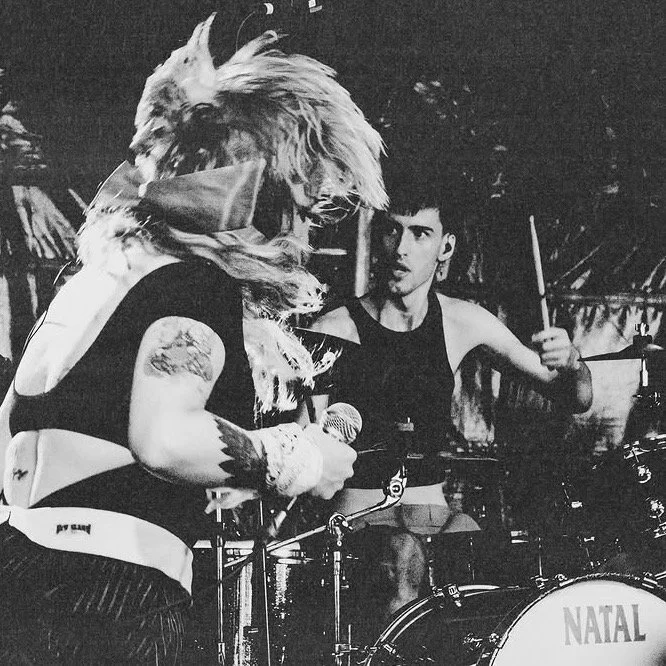 Black and white photo of two musicians on stage, one with a lion mask and the other playing drums, with the word 'NATAL' on the drum set.